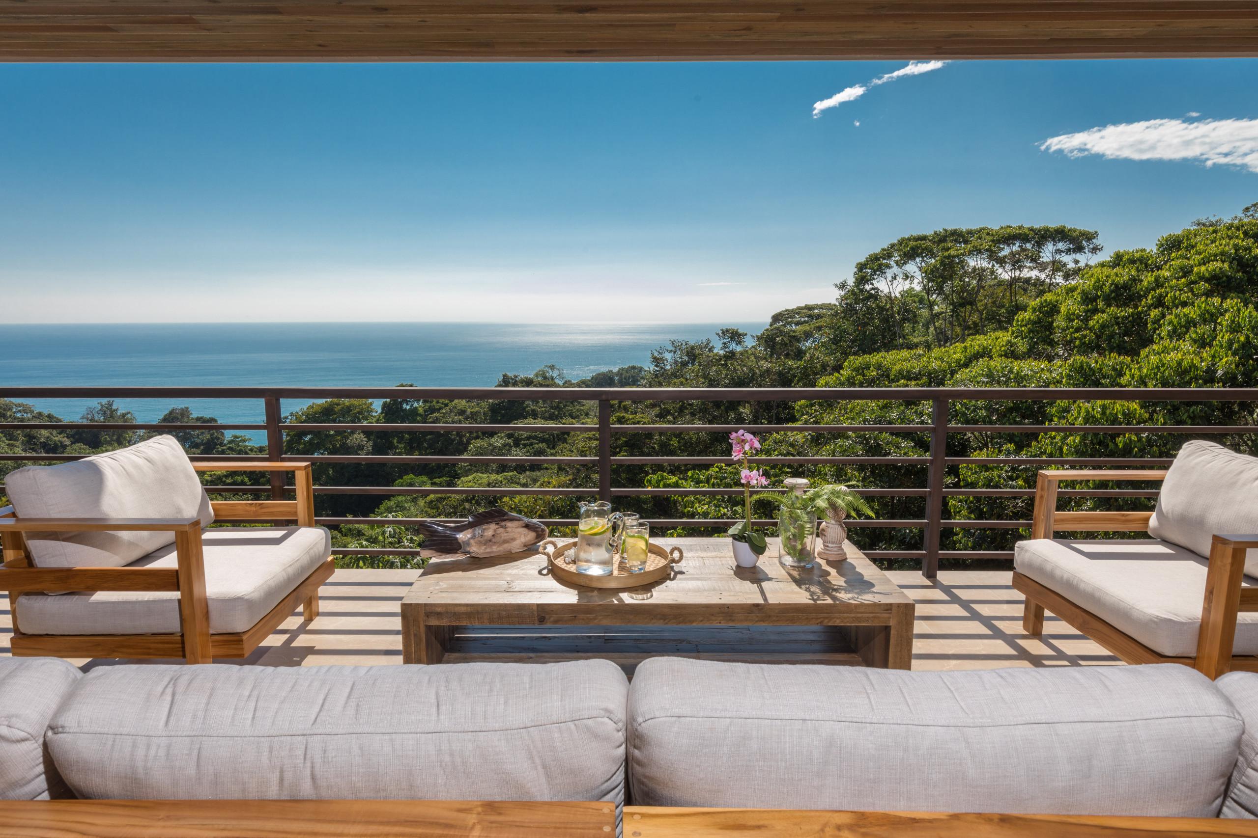 A modern outdoor living space, lit by natural light, presents a broad vista of the Pacific Ocean and lush jungle from a comfortable seating area on the terrace.