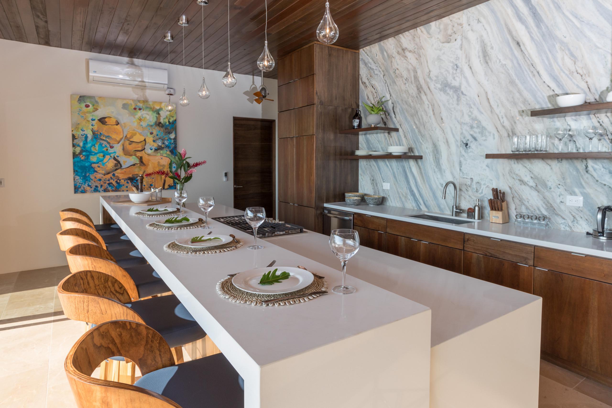 The kitchen space features a long island with six dining seats, wooden cabinetry, a wood-paneled ceiling, and a large veined marble wall, receiving natural light.