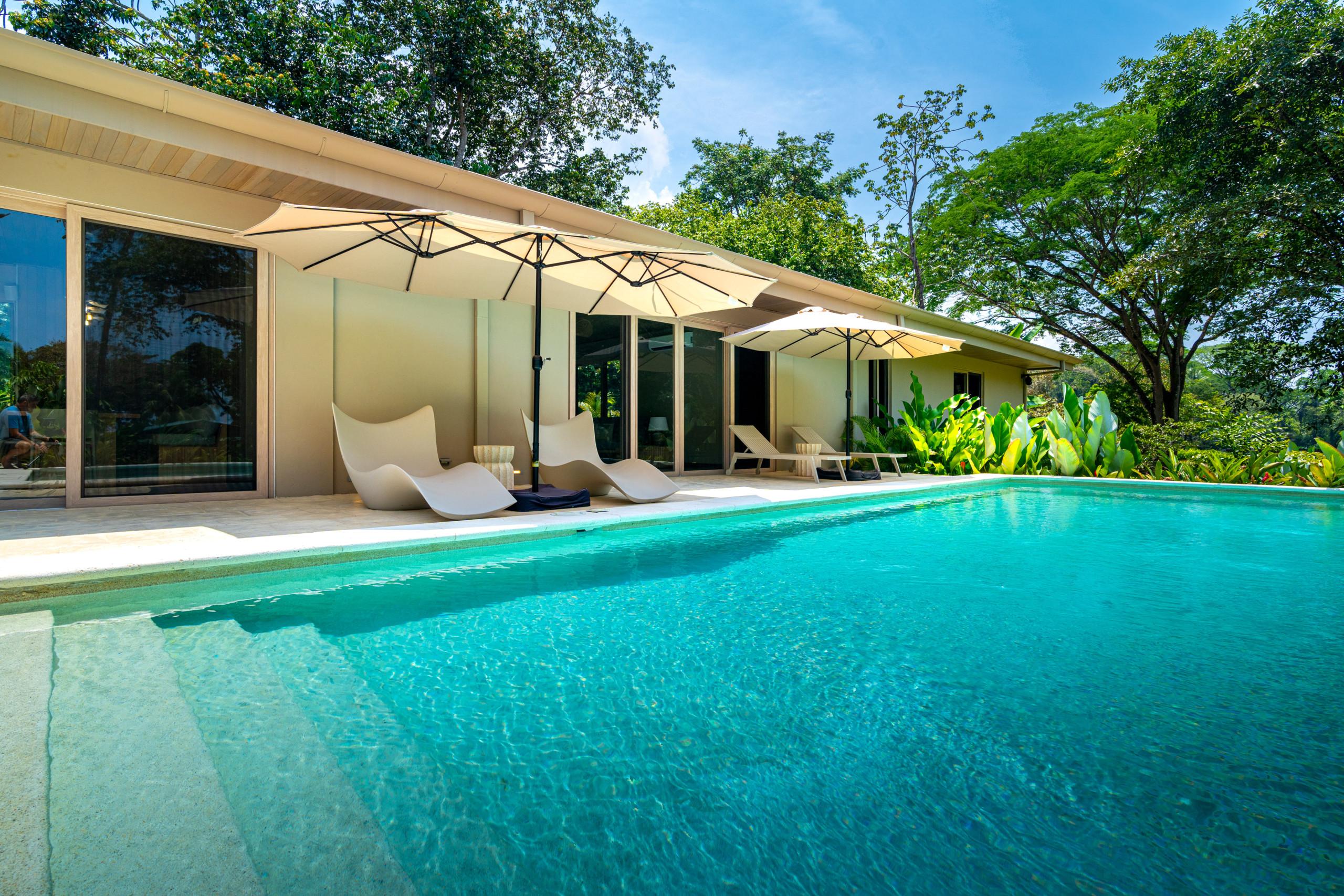 A private outdoor pool is adjoined by a patio with sun loungers and umbrellas, situated next to the villa's large glass doors and surrounded by abundant tropical trees.