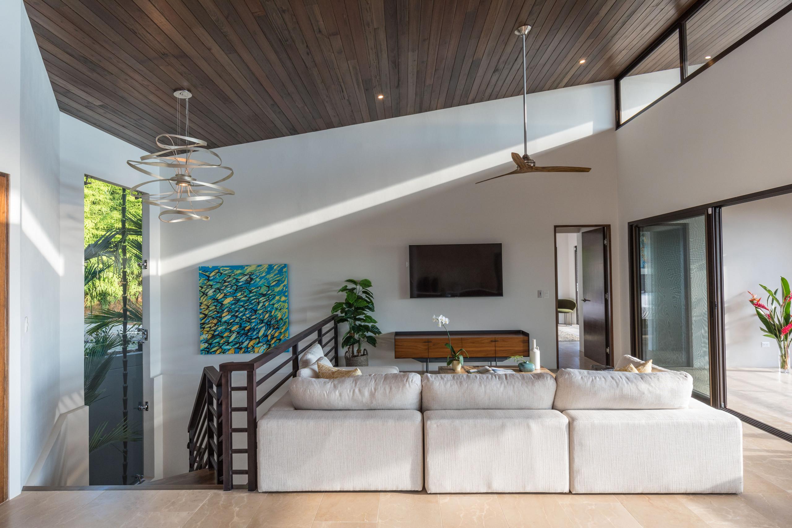 This bright, open living room features a high vaulted wooden ceiling, a large modular sofa, and natural light streaming through tall windows offering jungle views.