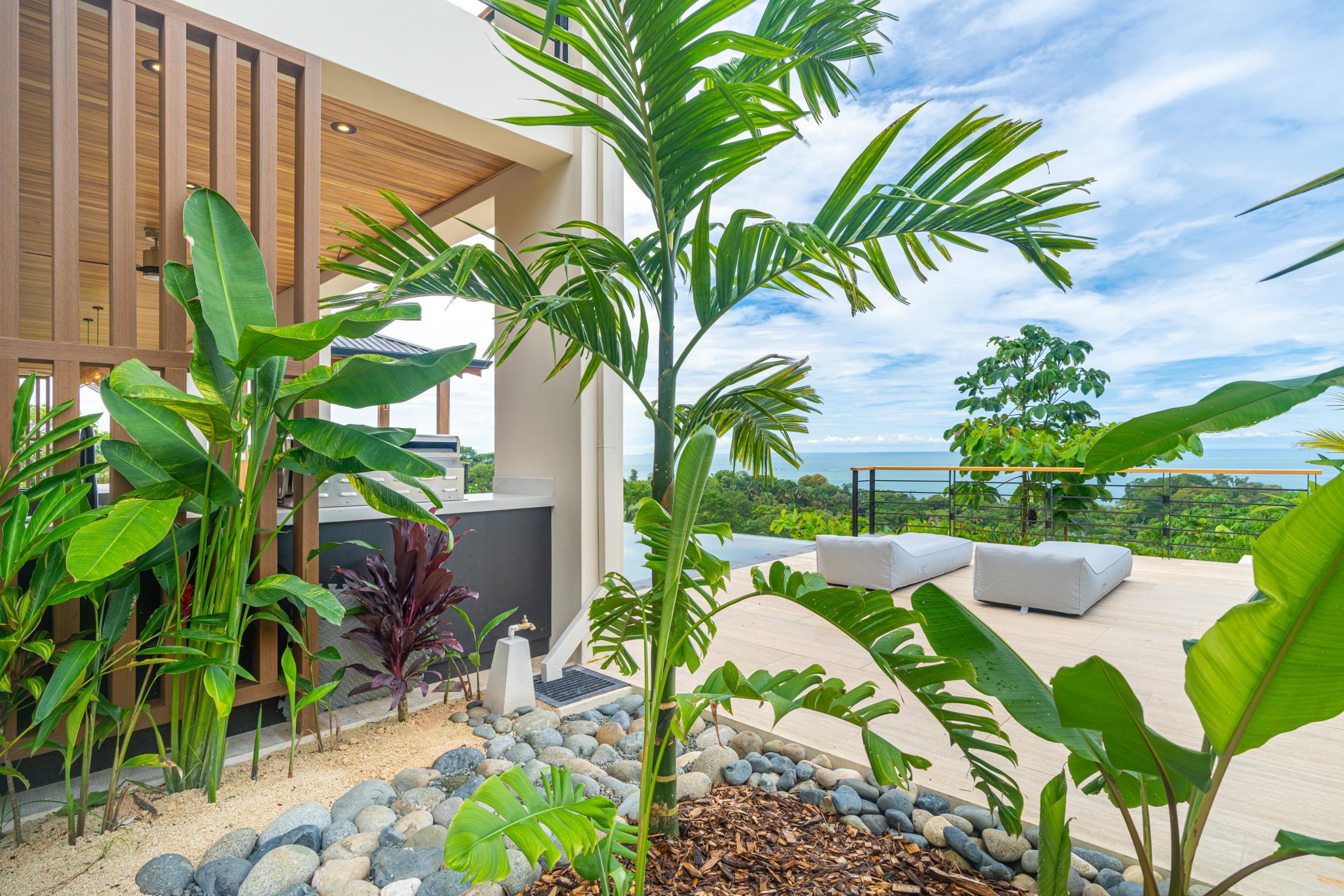 An outdoor terrace with an integrated kitchen and lounge seating is surrounded by tropical plants, framing views of an infinity pool, jungle, and the Pacific Ocean.