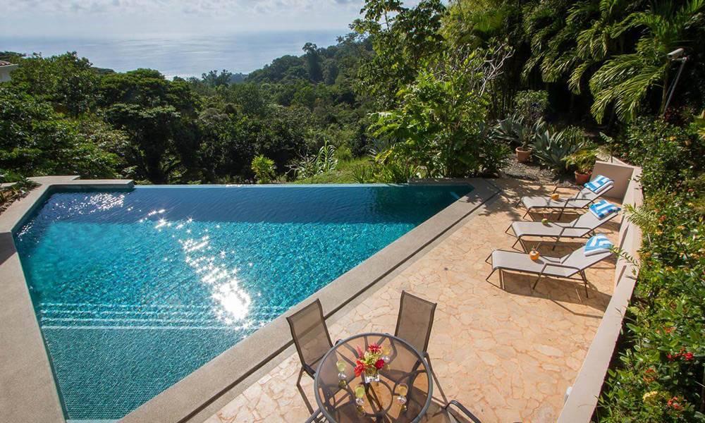 An infinity pool with sun-dappled water is set into a stone deck with lounge seating, offering views of the Pacific Ocean and surrounding jungle.