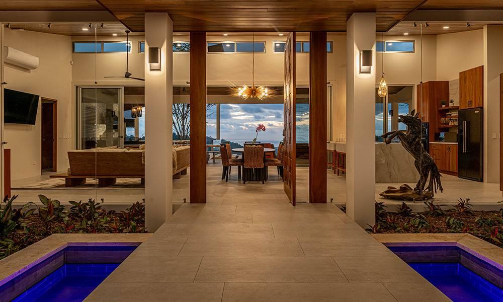 An outdoor entrance path with illuminated reflecting pools leads into a spacious, open living area showcasing panoramic ocean views.