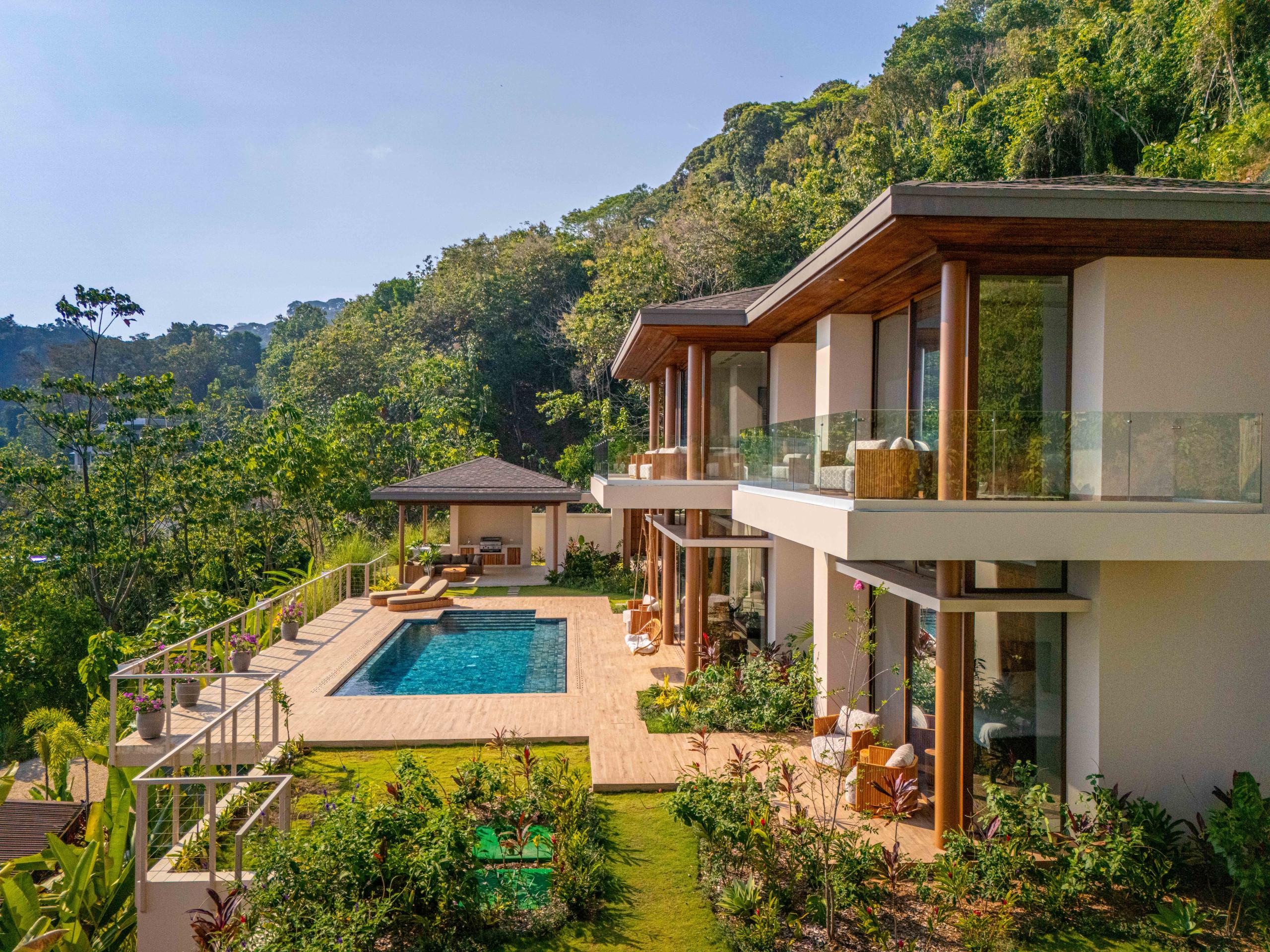 This multi-level villa, featuring warm wood accents and extensive glass, offers private balconies, an infinity pool with sun loungers, and an outdoor kitchen pavilion, all set within a lush tropical rainforest.