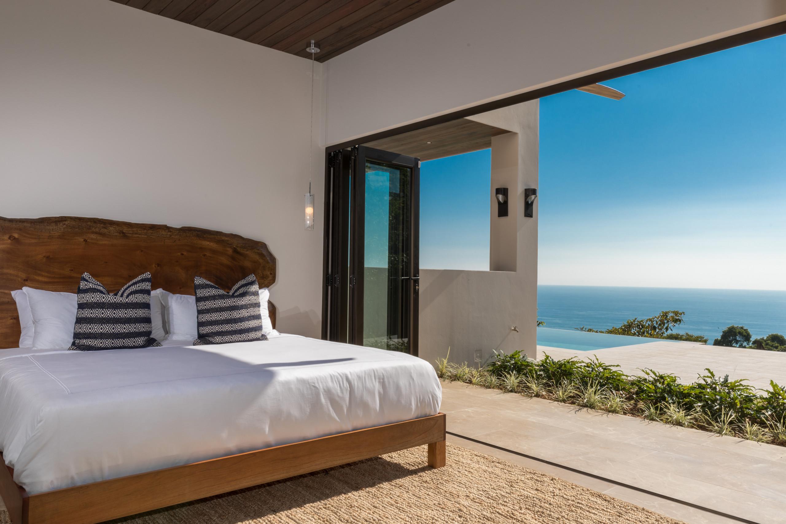 A king bedroom with a live-edge wooden headboard opens through bifold glass doors directly to a private terrace, inviting natural light and jungle-ocean views with an infinity pool.