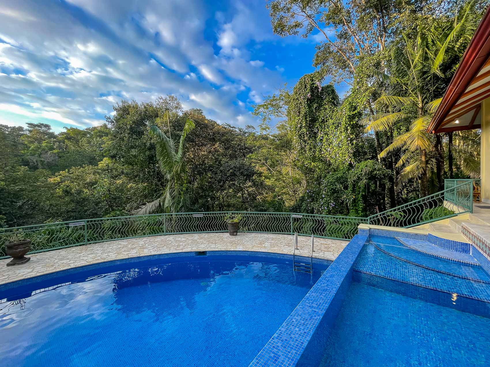 An outdoor tiled main pool and a connected wade pool are set on a terrace with a railing, offering views of the dense tropical canopy against a blue sky.
