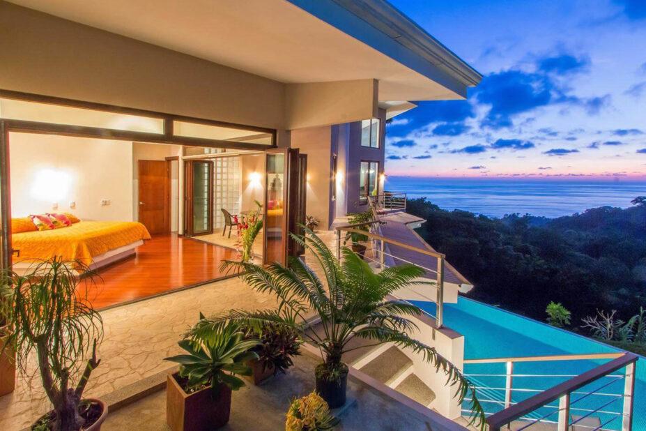 A bedroom with glass walls fully opens onto a private wrap-around balcony, revealing sweeping ocean and jungle views and an infinity pool under a vibrant sunset.