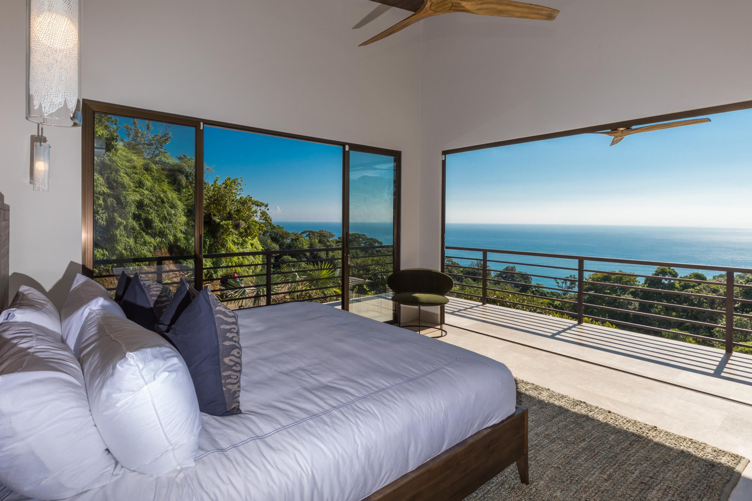 A bright bedroom with a king-size bed opens through expansive windows to a terrace, offering panoramic ocean and jungle views.