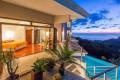 A private terrace connects to a glass-walled bedroom, offering unobstructed views of an infinity pool and the Pacific Ocean at dusk.