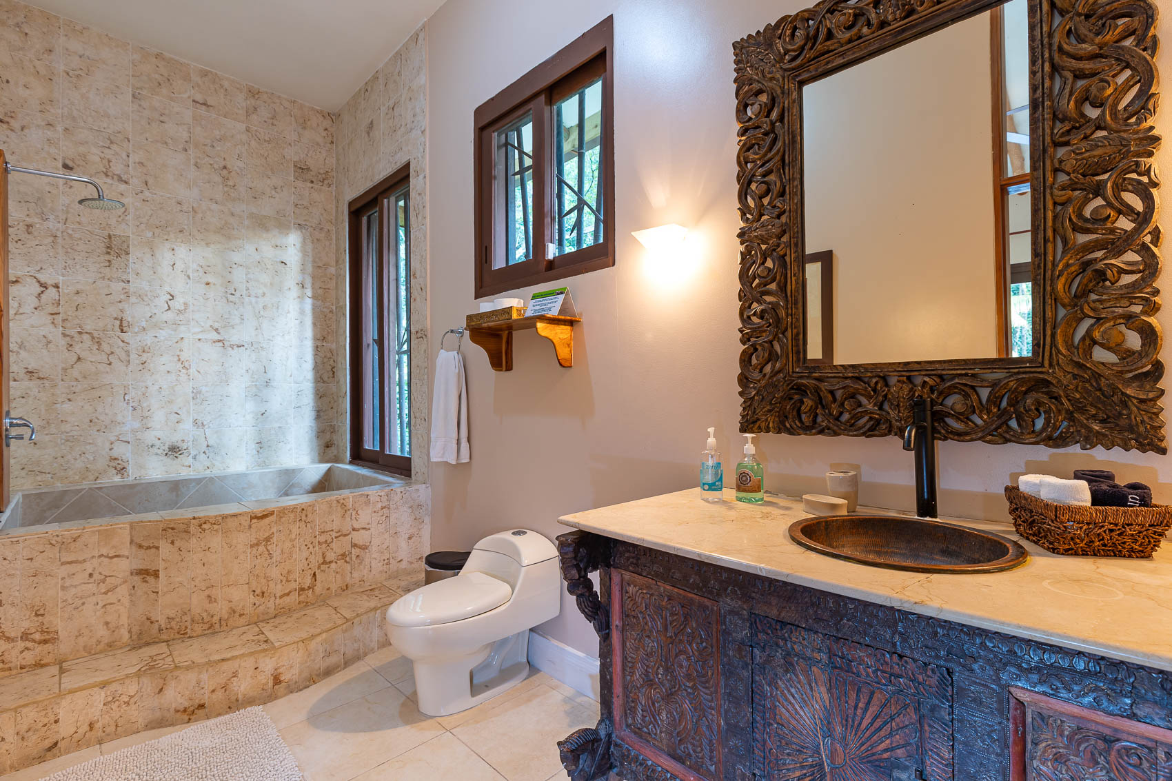 This bathroom features a deep, tiled bathtub/shower, a hand-carved vanity with a unique basin under an ornate mirror, and windows offering natural light and views of the lush surroundings.