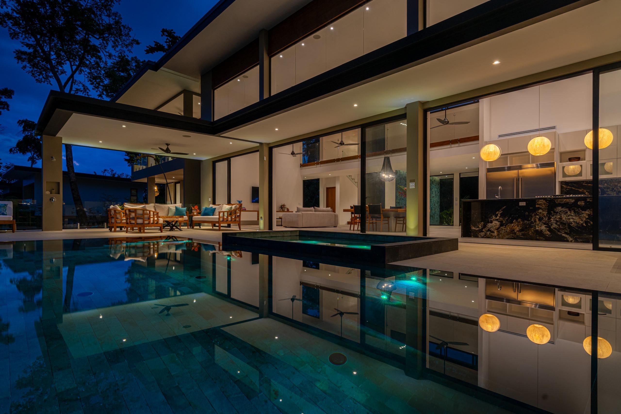 The illuminated modern villa at dusk showcases an expansive pool reflecting the open-plan living areas, dining room, and kitchen visible through floor-to-ceiling glass walls.