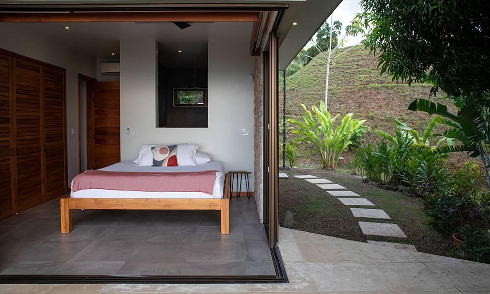 A bedroom with a king bed features bi-fold doors opening directly to a garden pathway and lush hillside, providing natural light and an outdoor connection.