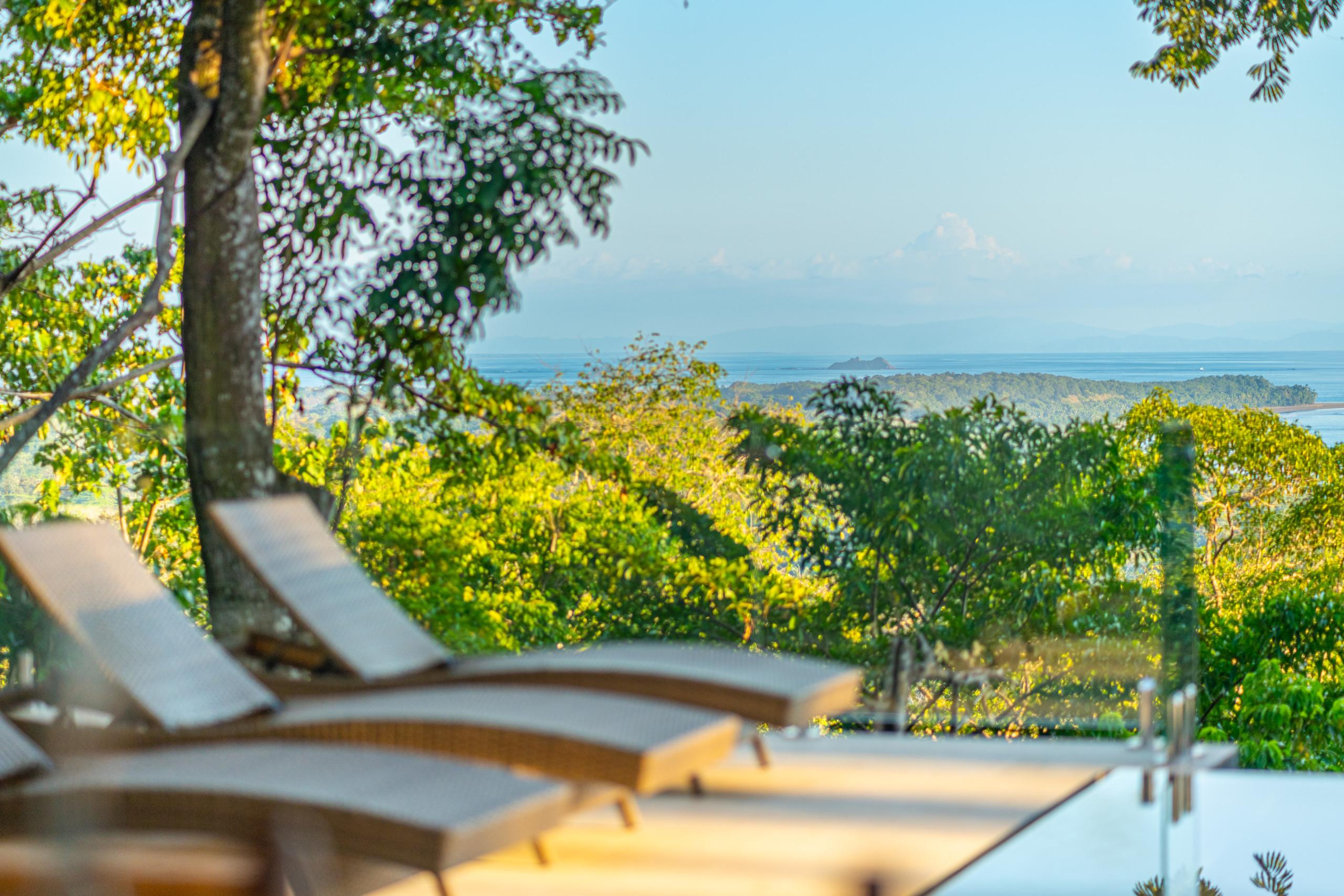 The sunlit pool deck with lounge chairs presents a wide view of the tropical forest and the sea stretching to the horizon.