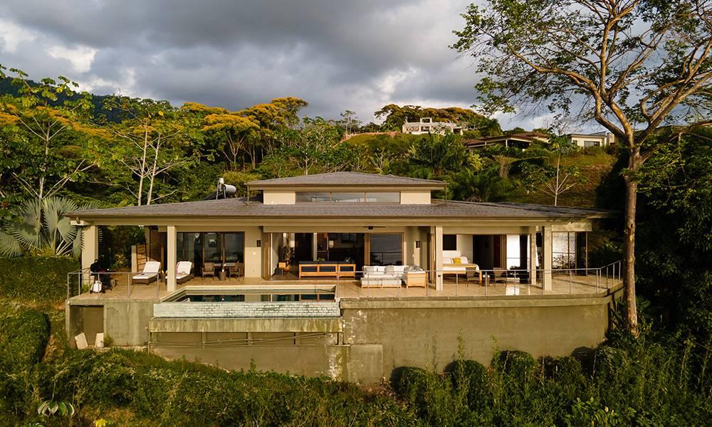 The single-level villa features a broad covered terrace with a visible pool, outdoor lounging, and expansive glass doors opening to a lush tropical setting.