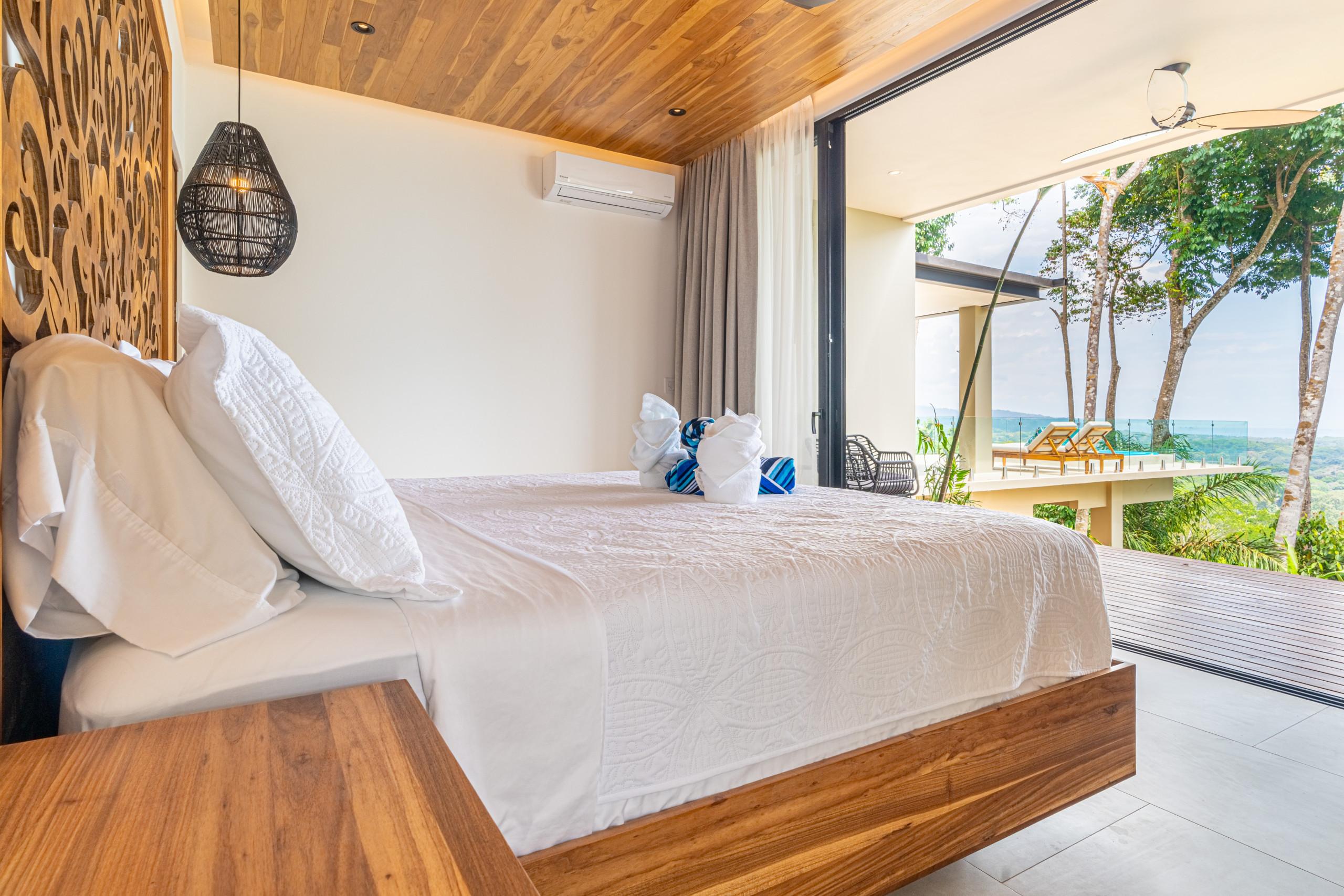 A bedroom featuring a king bed with a carved wooden headboard and a wood-paneled ceiling, bathed in natural light from sliding glass doors that lead to a private terrace overlooking the ocean.