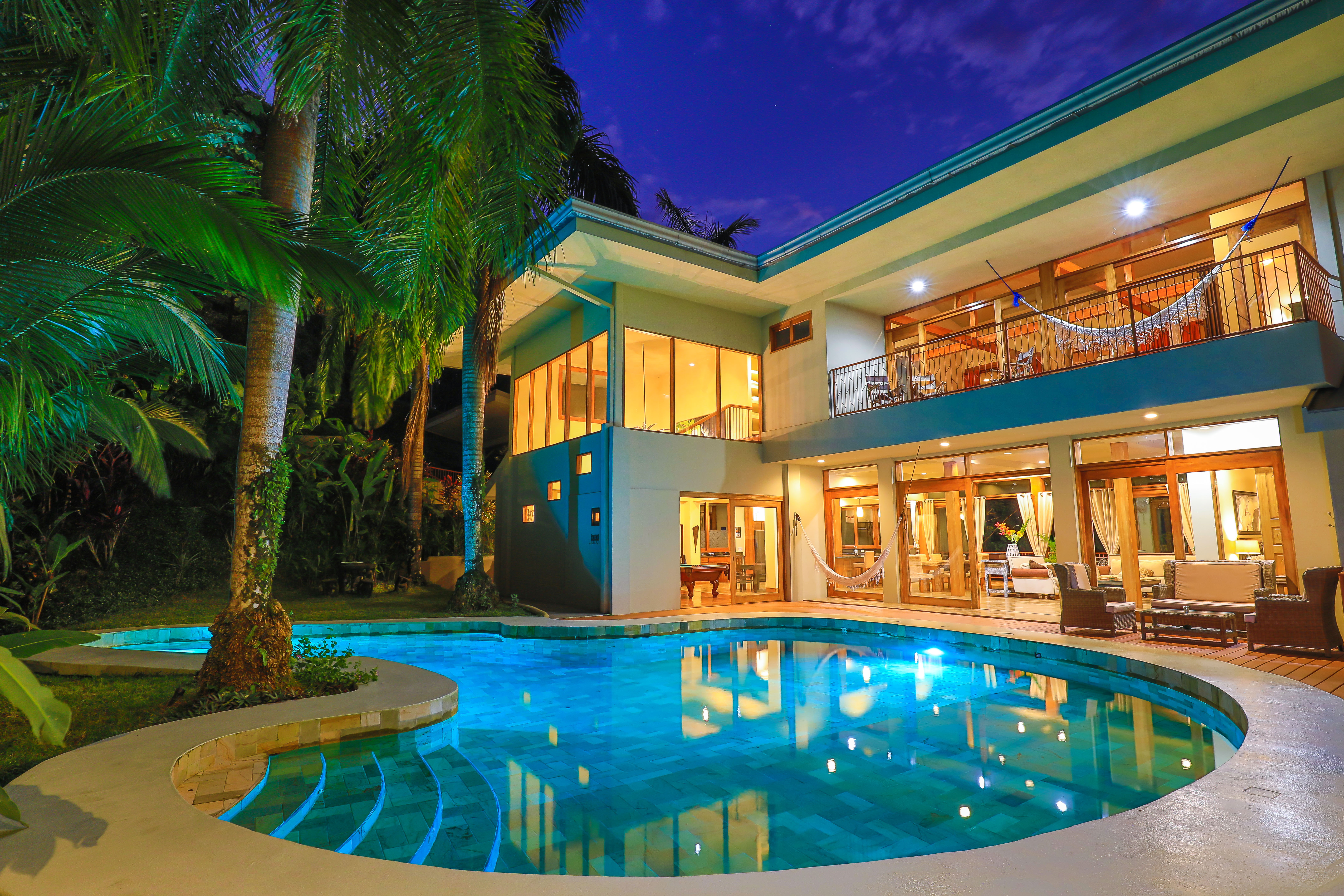 The multi-level villa illuminates its interior spaces through expansive windows and glass doors, opening directly to a large swimming pool and deck amidst tropical foliage under the evening sky.