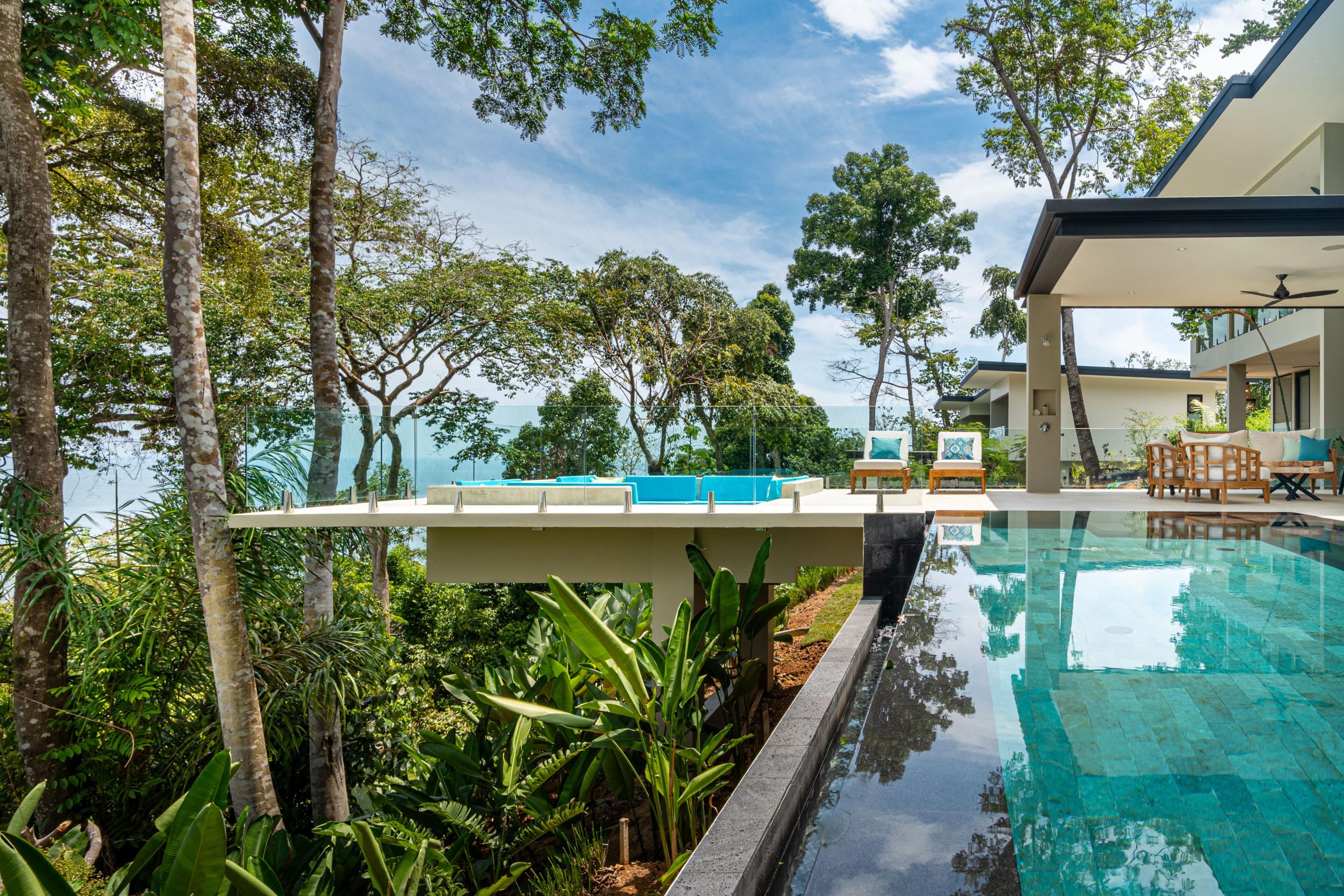 An infinity pool with turquoise tiling connects to a raised sun deck with a jacuzzi, overlooking the ocean and tropical forest.