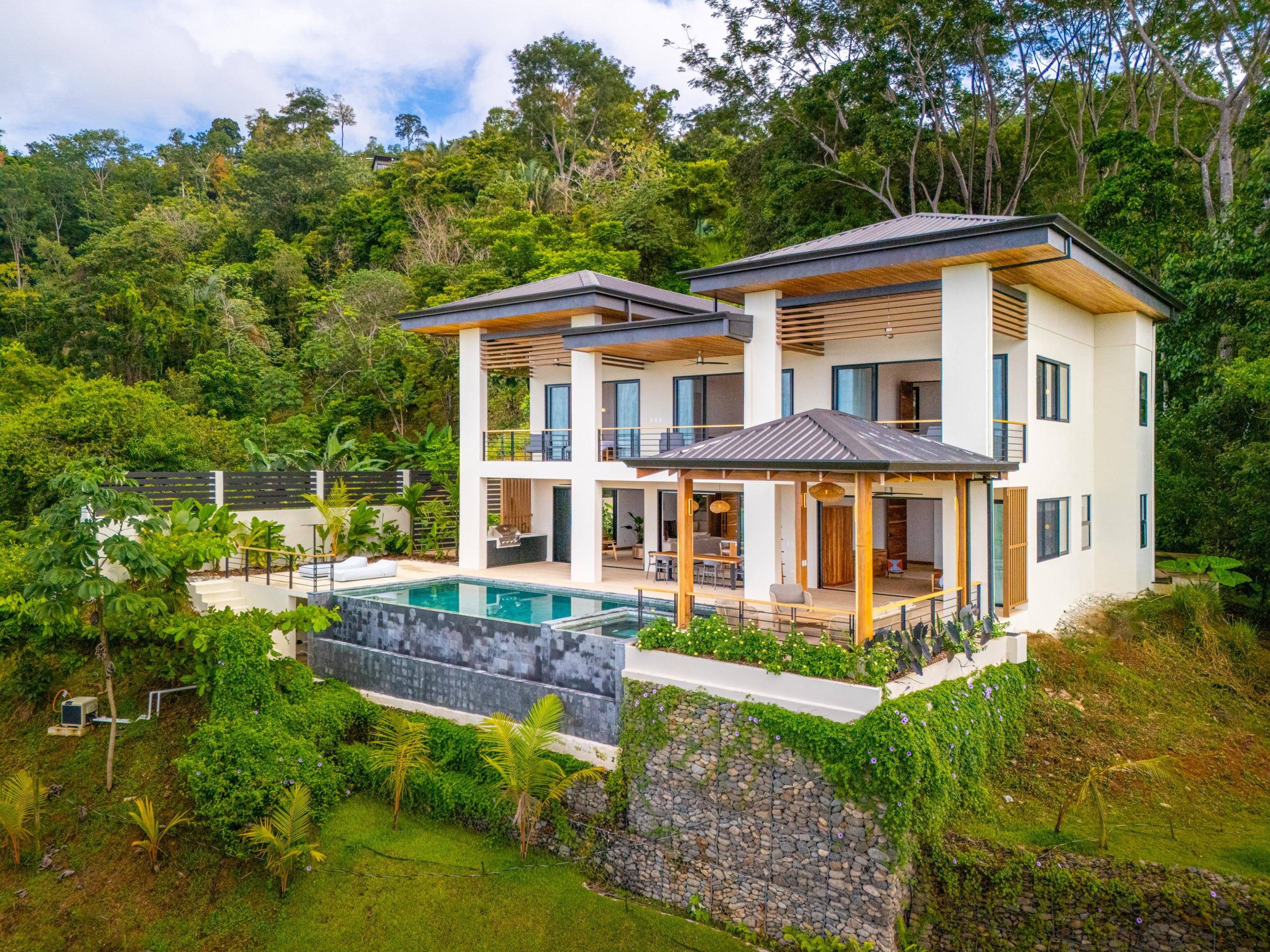 A modern two-story villa featuring white facades, wooden accents, multi-level terraces, an infinity pool with an adjacent jacuzzi, and a covered palapa, all set against a dense tropical jungle hillside.
