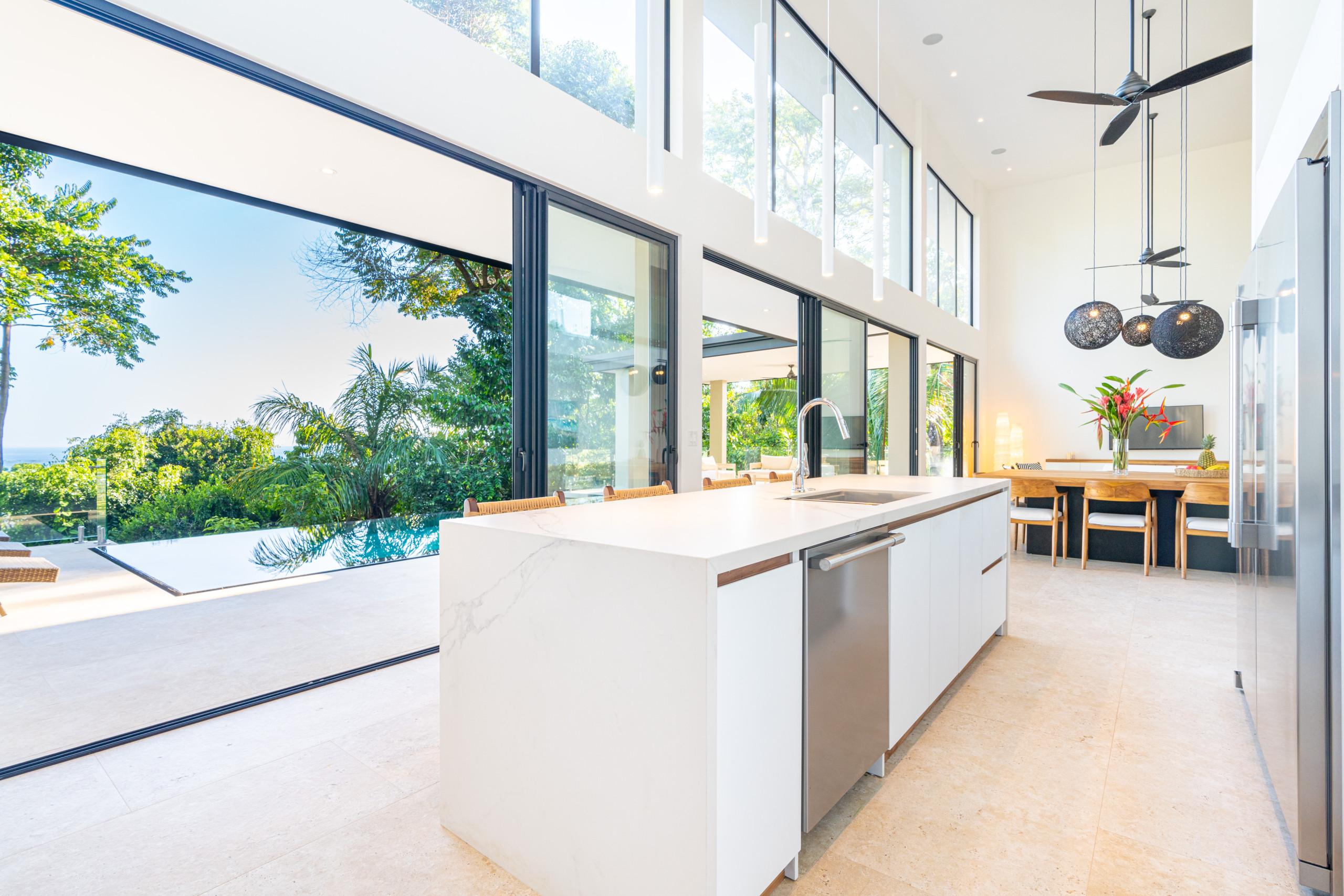 The high-ceilinged kitchen with a large island and adjacent dining area receives abundant natural light and opens through expansive glass doors to an infinity pool and tropical ocean views.