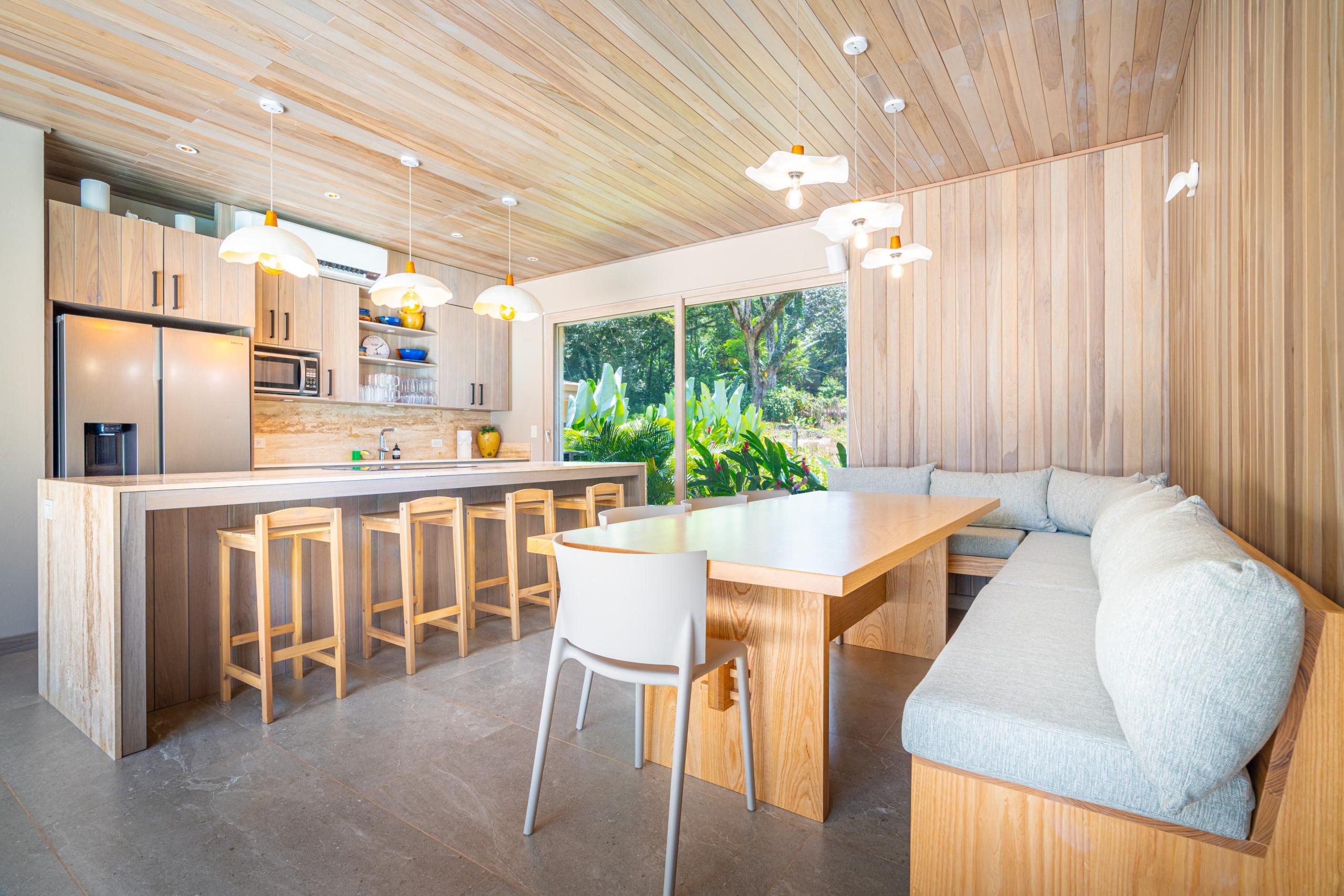A bright open-plan dining area with an integrated kitchen, featuring warm wood-paneled walls and ceiling, an L-shaped banquette, and expansive windows offering views of lush tropical greenery.