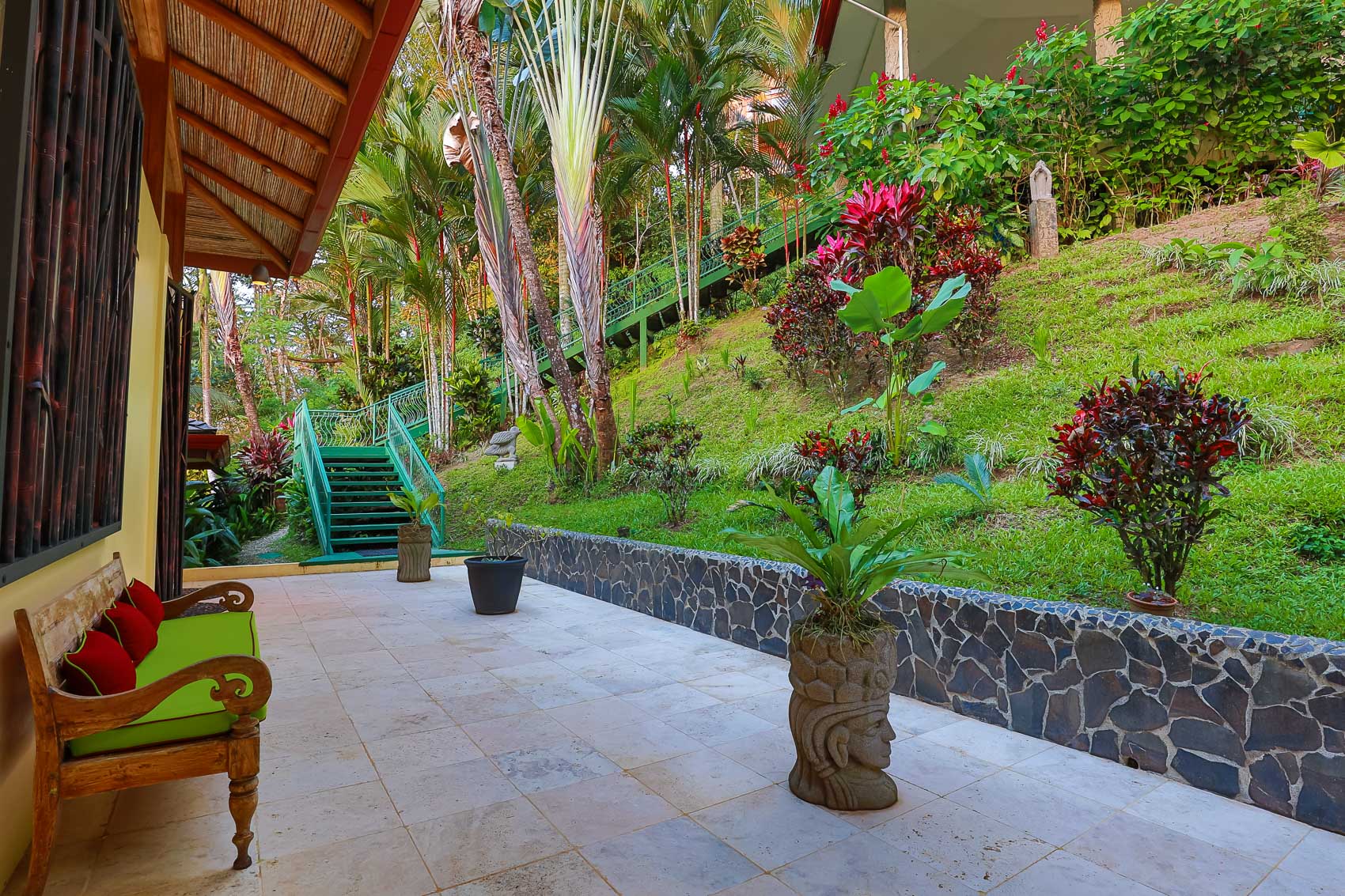 This outdoor terrace, partially covered by a bamboo roof, features a paved floor and a wooden bench, opening to a tropical hillside garden with a winding green staircase.