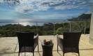 An open outdoor terrace with a glass railing provides an elevated seating area directly overlooking the ocean and coastline under abundant natural light.