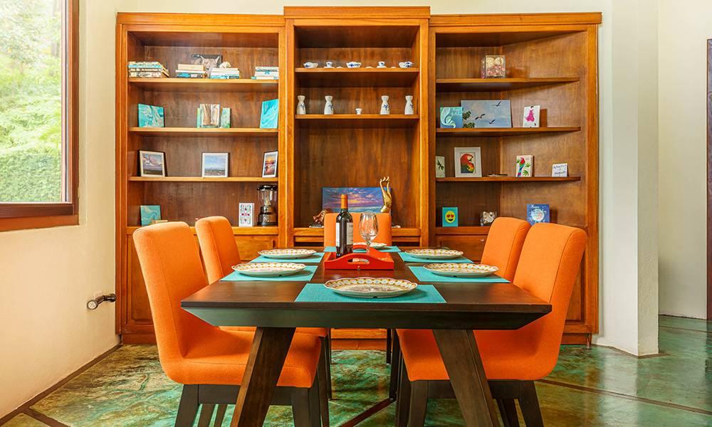This dining area features a dark wood table with six vibrant orange chairs, a large built-in wooden bookshelf, and natural light from a window looking out to tropical greenery.