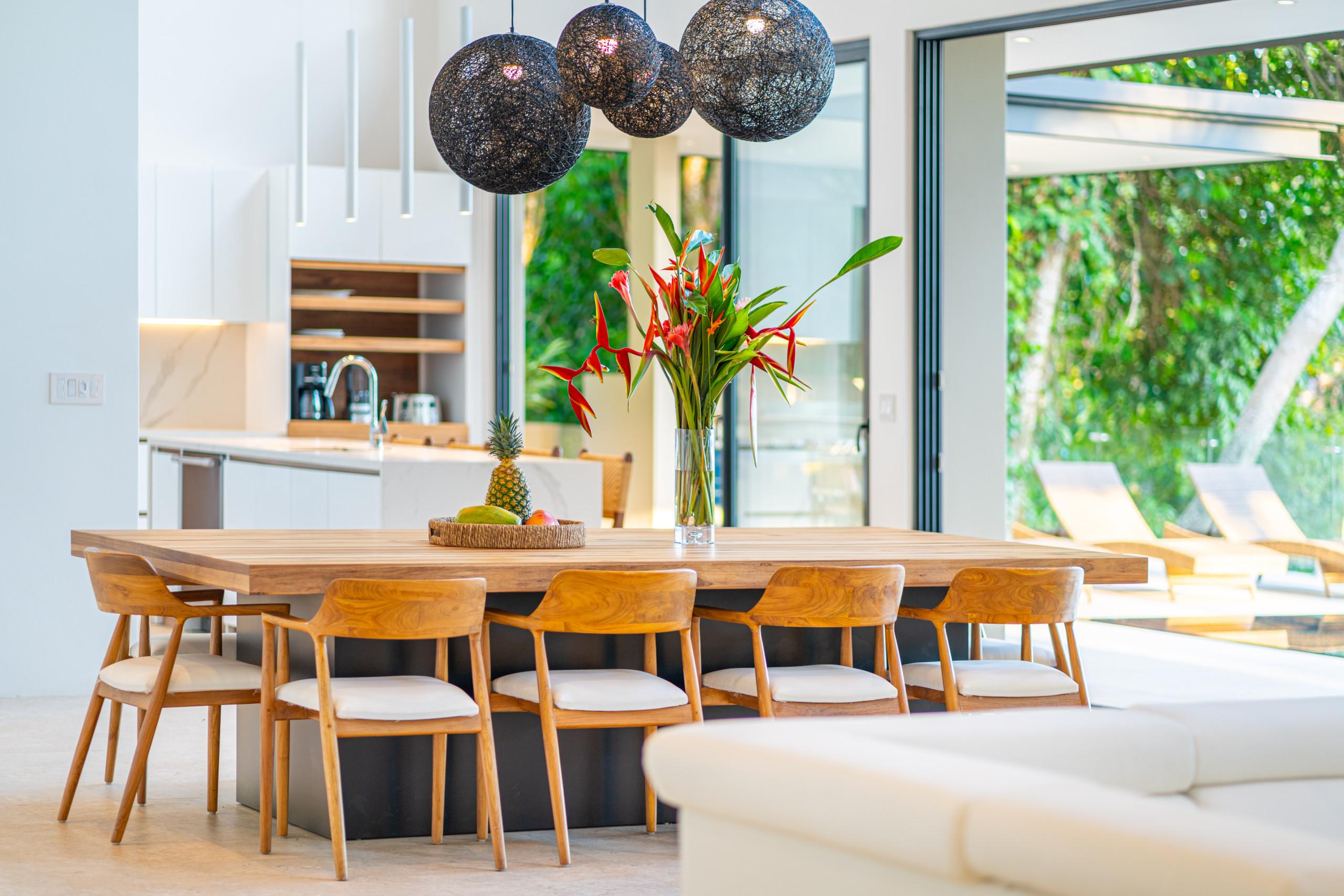 A modern dining space with a large wooden table and seating for eight connects to a contemporary kitchen, opening via glass doors to a sunlit outdoor terrace with tropical views.