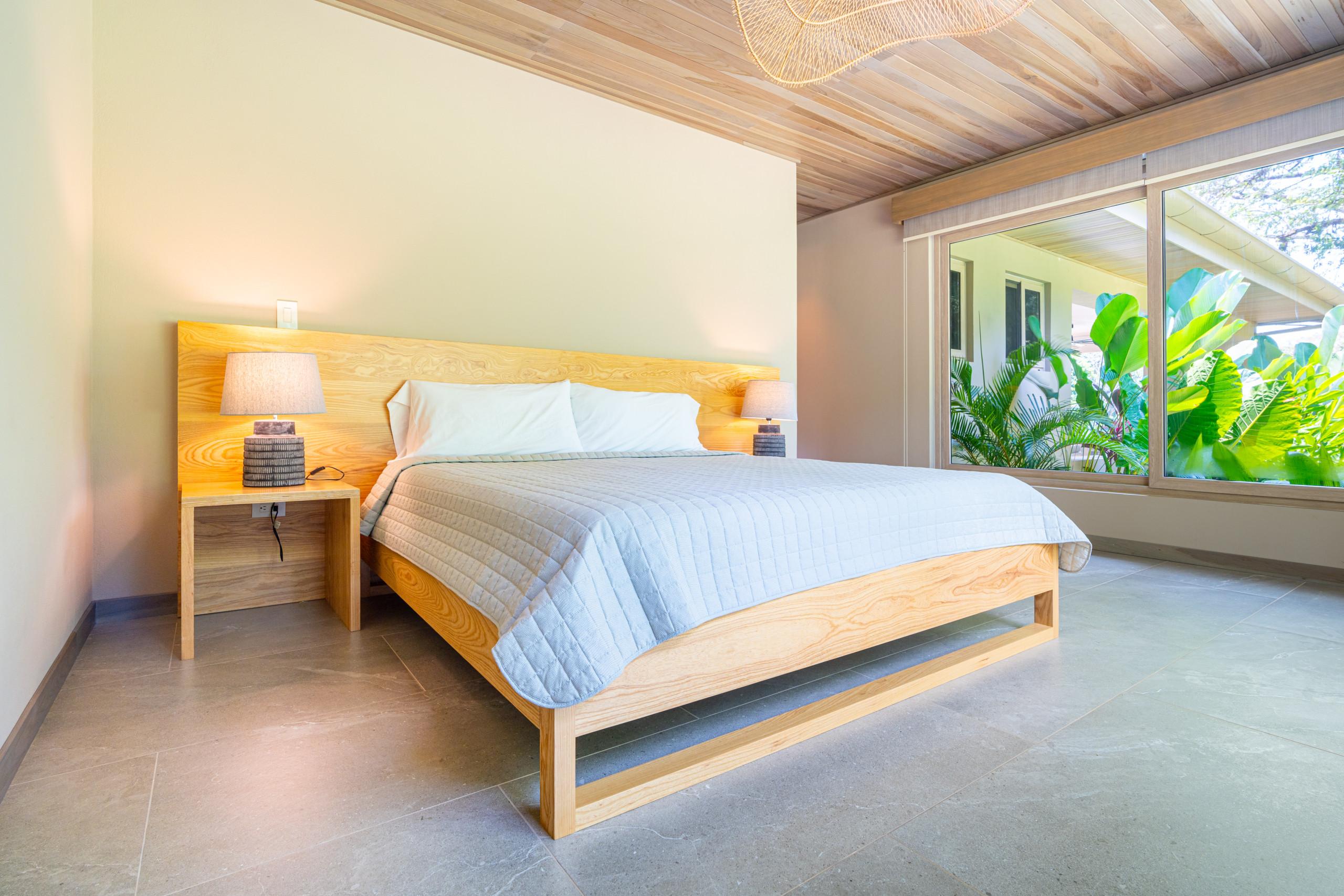 A bright bedroom features a wooden bed frame and headboard, light walls, a slatted wood ceiling, and expansive windows offering views of lush tropical greenery.