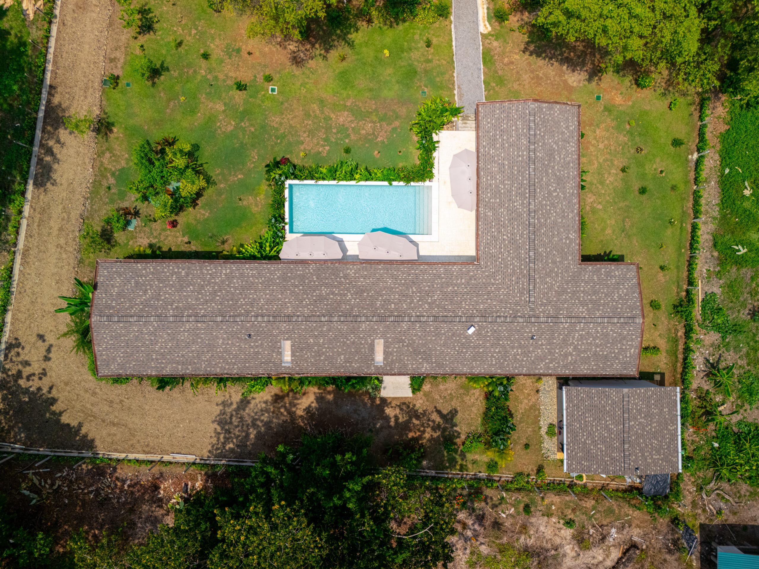 An aerial view presents the L-shaped villa, a private rectangular pool with a paved terrace and umbrellas, surrounded by extensive green gardens that border dense tropical foliage.