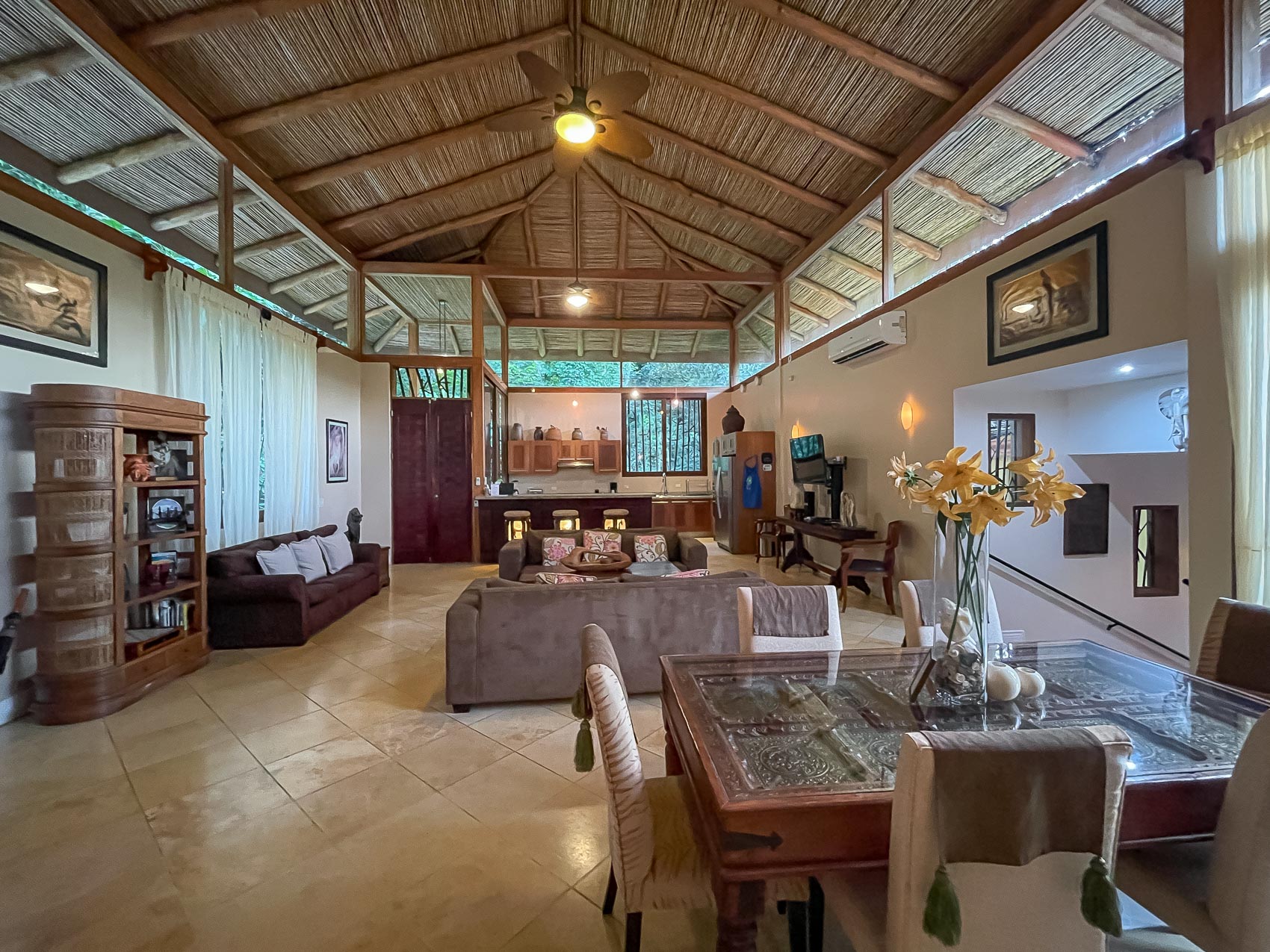 A spacious open-concept living and dining area features a high vaulted bamboo ceiling, natural light from clerestory windows, and views of lush greenery.