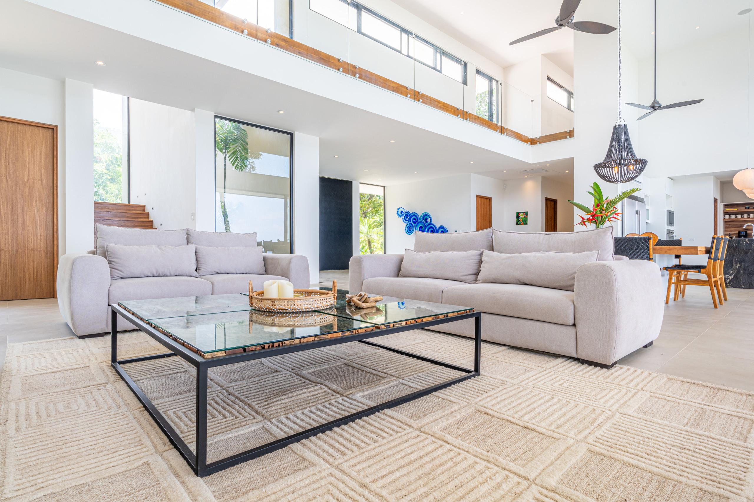 This double-height living room features large windows with views of tropical greenery, abundant natural light, and an open connection to the dining and kitchen areas, overlooked by a second-story walkway.