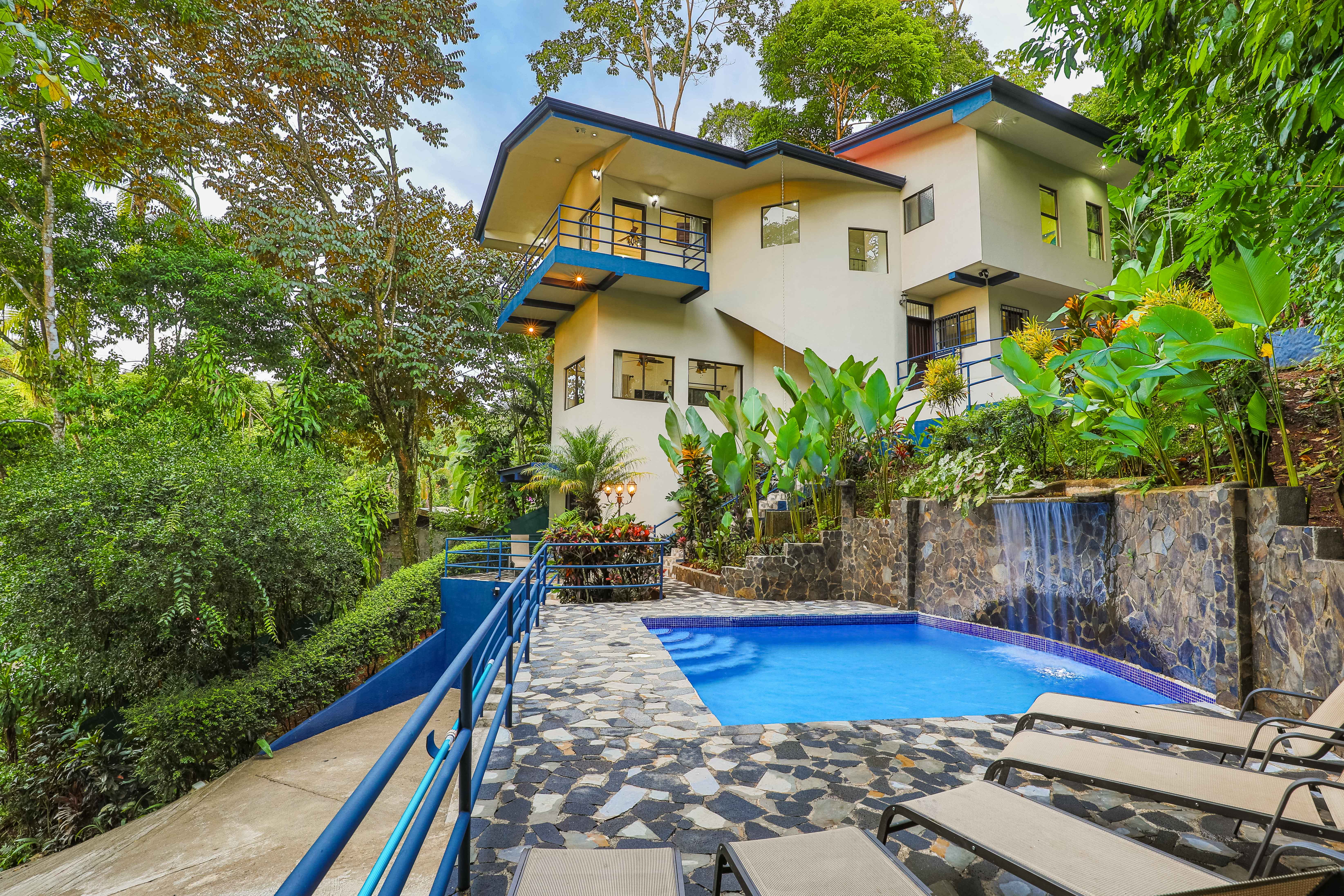 A contemporary multi-level villa with blue accents, multiple balconies, and large windows overlooks a private pool with a waterfall, all set within a vibrant tropical rainforest.
