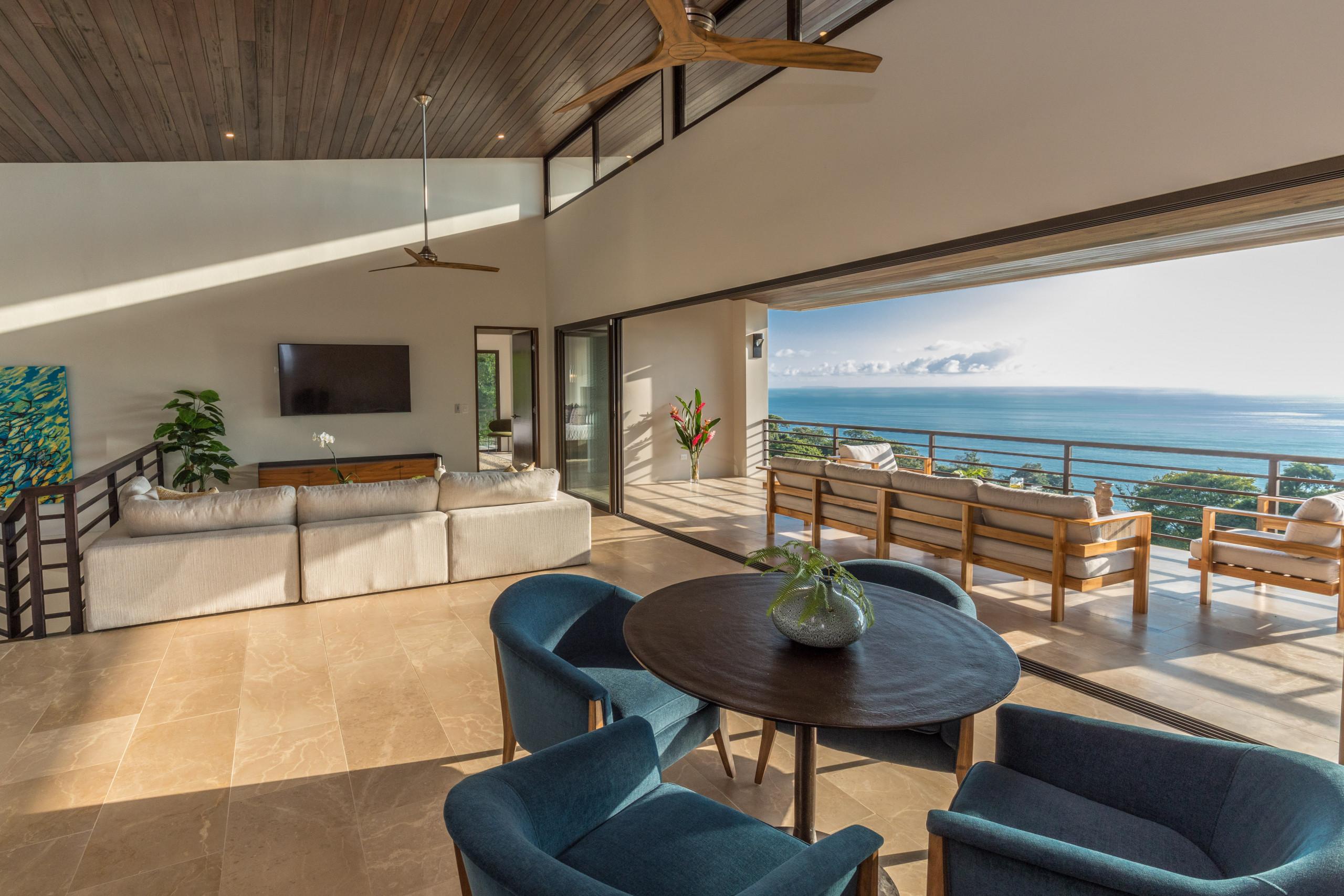 An open living room, featuring a wood-paneled ceiling and tiled floor, flows onto an outdoor terrace with an expansive ocean view and abundant natural light.