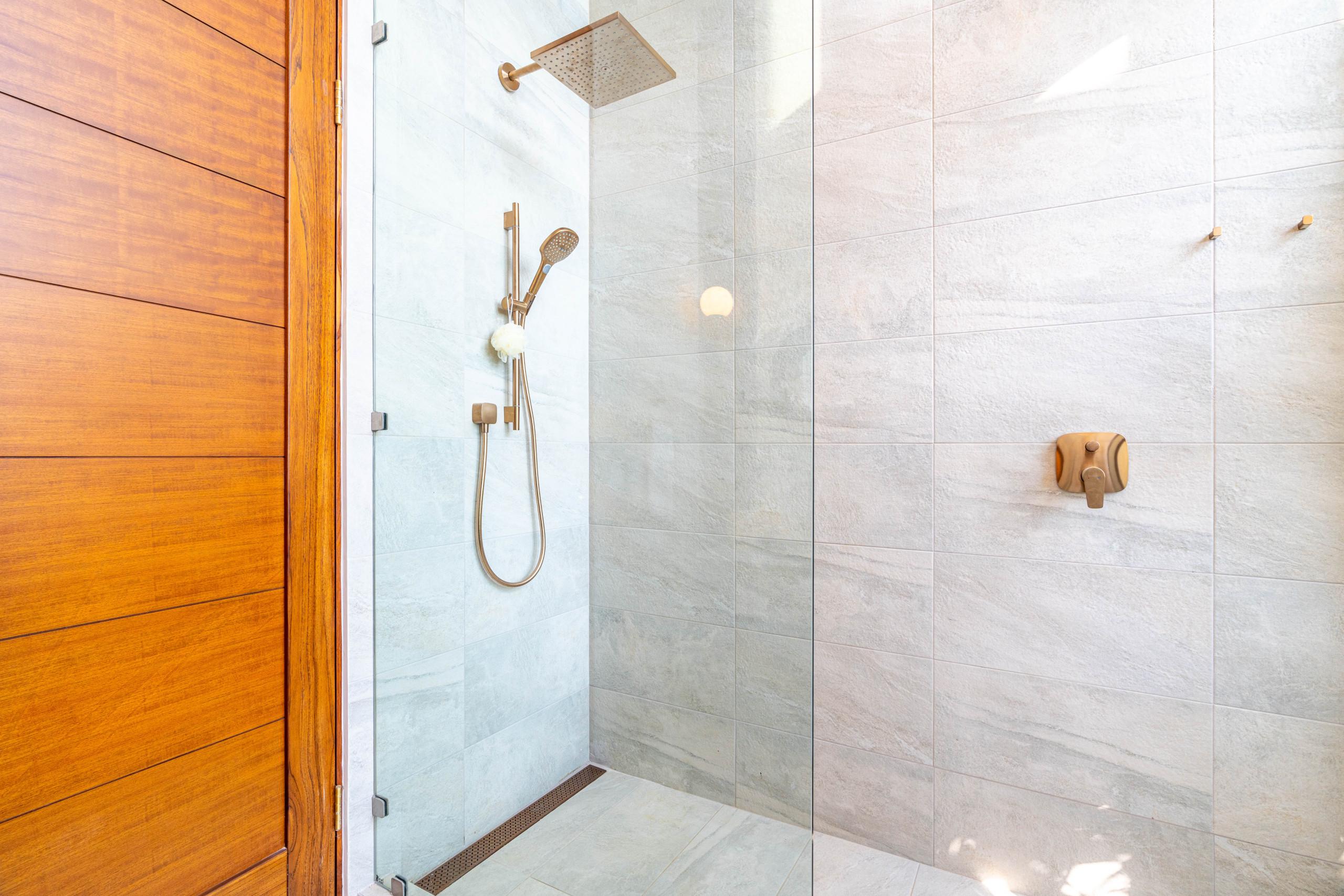 This bathroom features a glass-enclosed rainfall shower with light stone-look tiles and bronze fixtures, beside a warm wooden door, illuminated by ambient light.