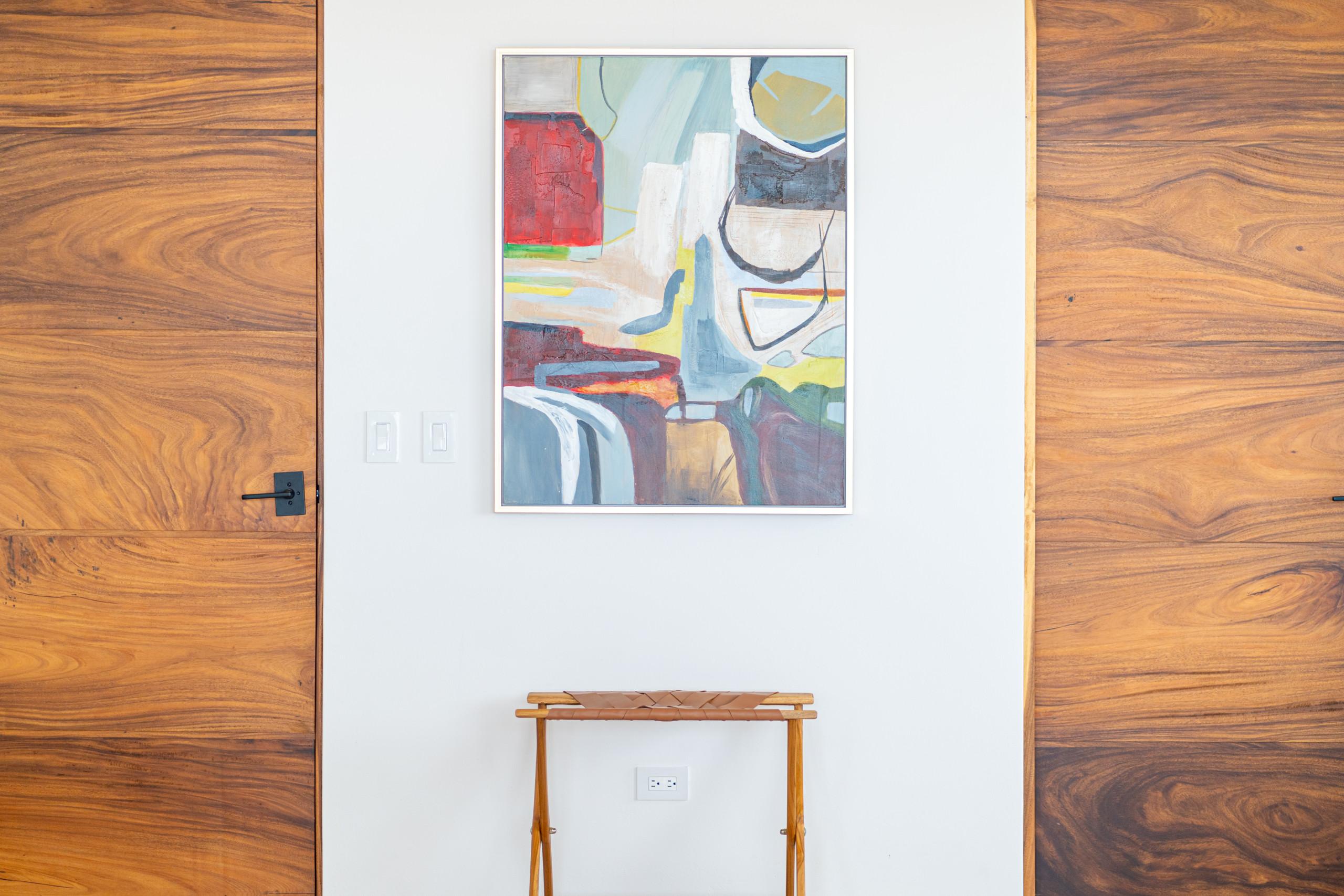Natural wood doors with distinctive grain frame a white wall featuring abstract art and a woven leather luggage rack, adding a warm, contemporary accent to the interior.