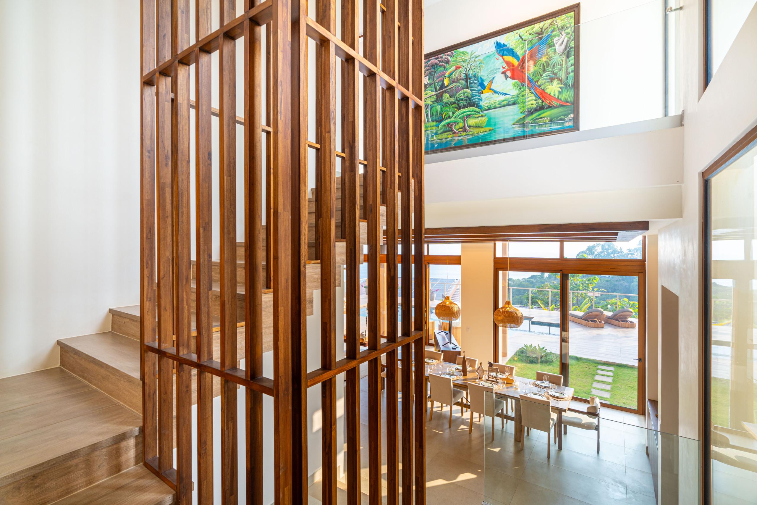 This bright, open-plan space features a prominent wooden slatted staircase, a dining area with a long wooden table, and expansive glass doors opening to a terrace with jungle and ocean views.