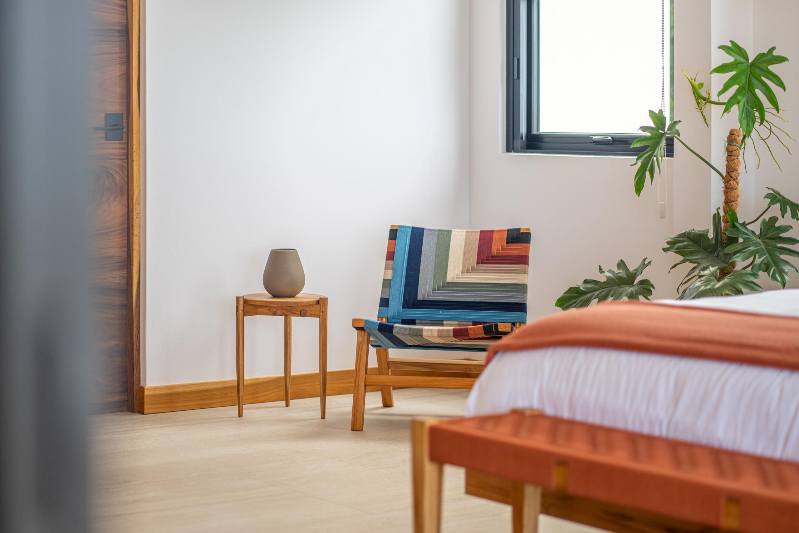 A bright bedroom features a comfortable bed, a colorful Masaya chair, a large window letting in natural light, and a wooden door.