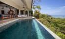 An infinity pool with dark tiling extends toward the ocean horizon, bordered by tropical foliage and positioned adjacent to a shaded open-air living space.
