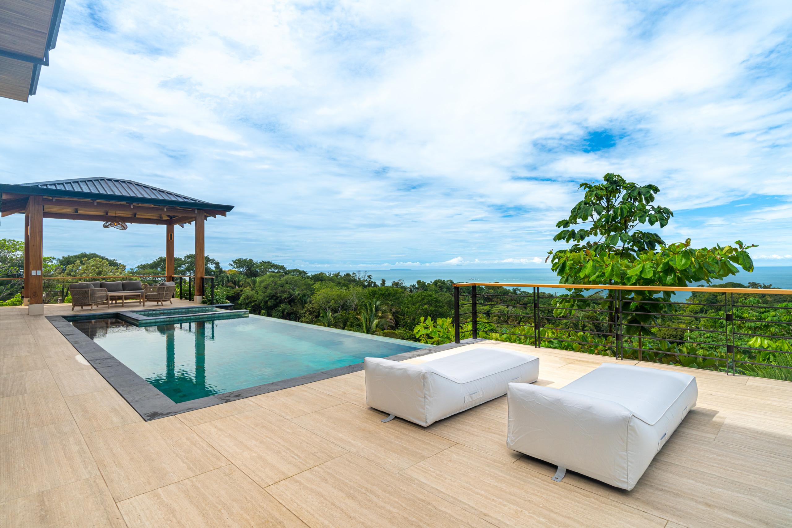 An infinity pool with an integrated jacuzzi is set on a tiled terrace featuring lounge seating and a shaded palapa, all overlooking a vast jungle leading to the ocean.