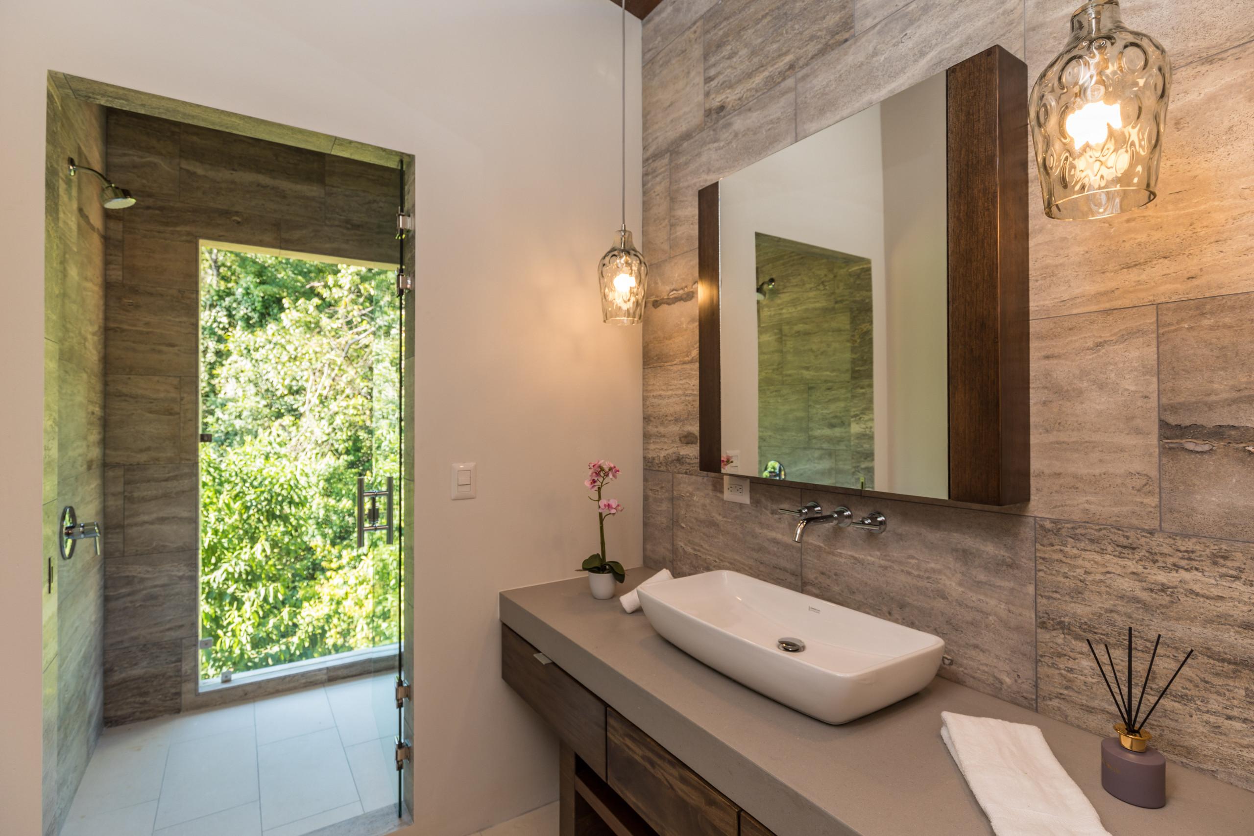 This contemporary bathroom integrates natural light and jungle views through an open shower design, featuring warm stone and wood finishes.