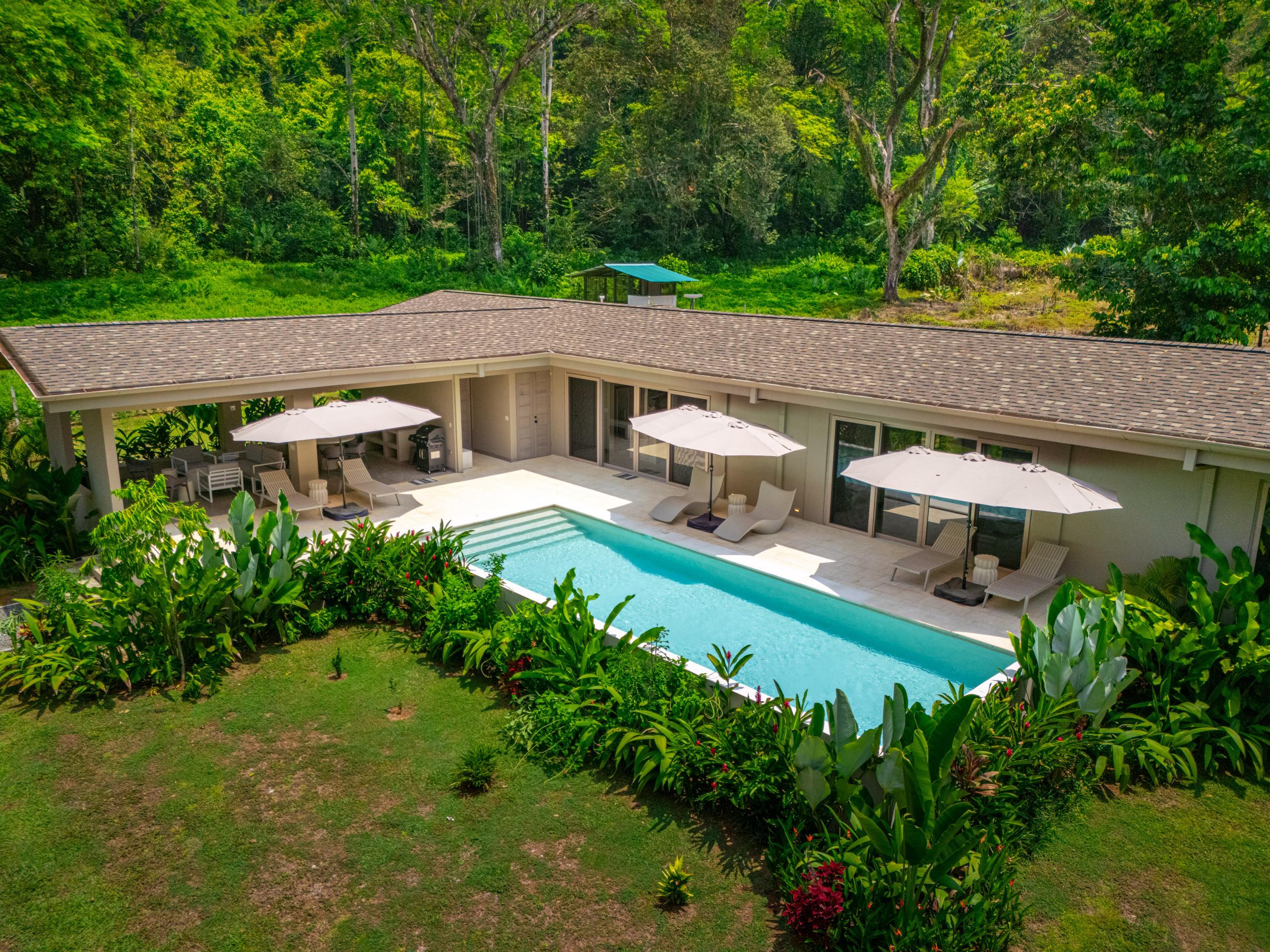 An L-shaped villa features a private pool, patio with sun loungers, and a covered outdoor dining and BBQ area, all bordered by manicured lawns and dense tropical jungle.