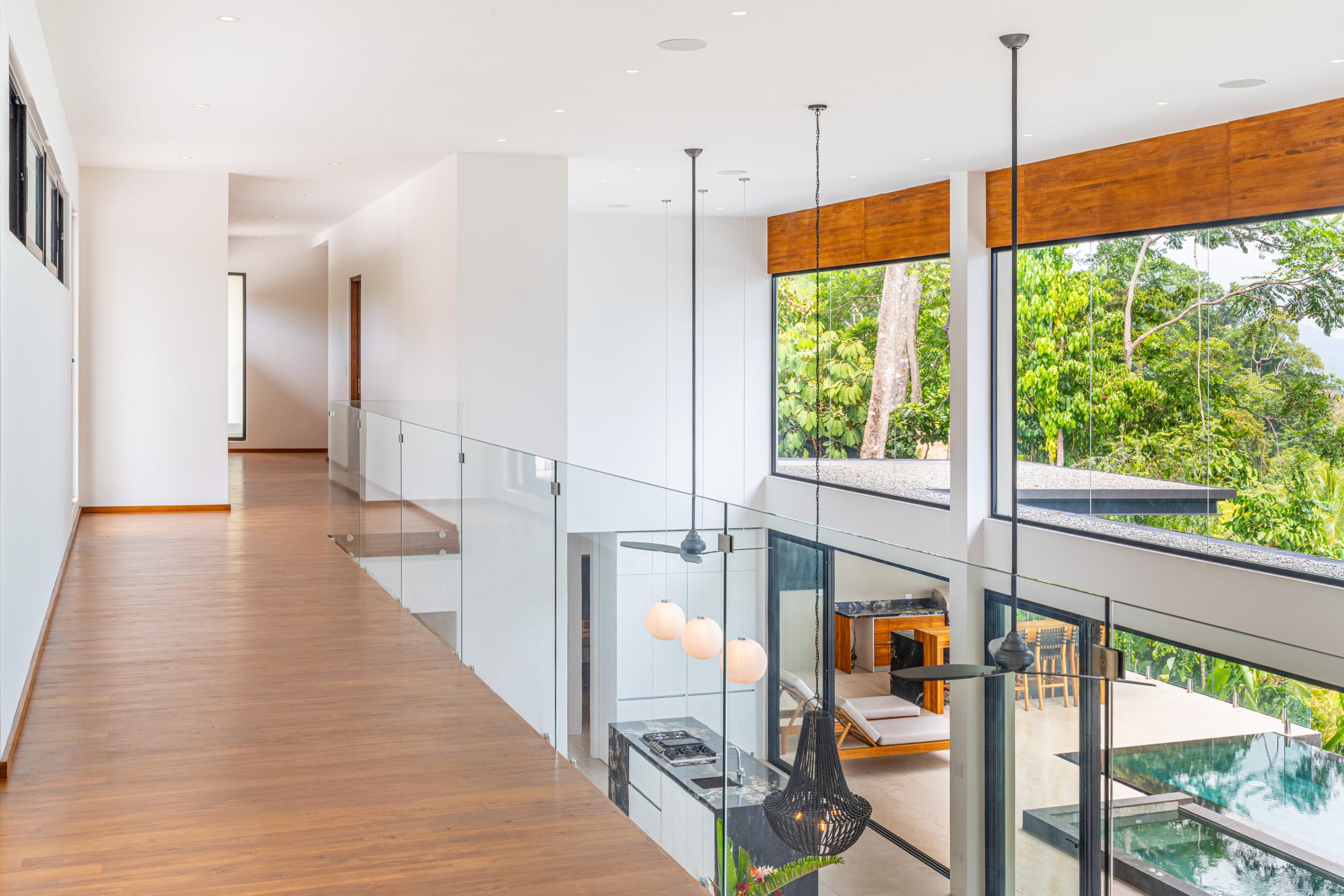 This bright, open multi-level interior provides an upper walkway with glass railings, overlooking a modern kitchen below and offering views of lush tropical jungle through large windows.