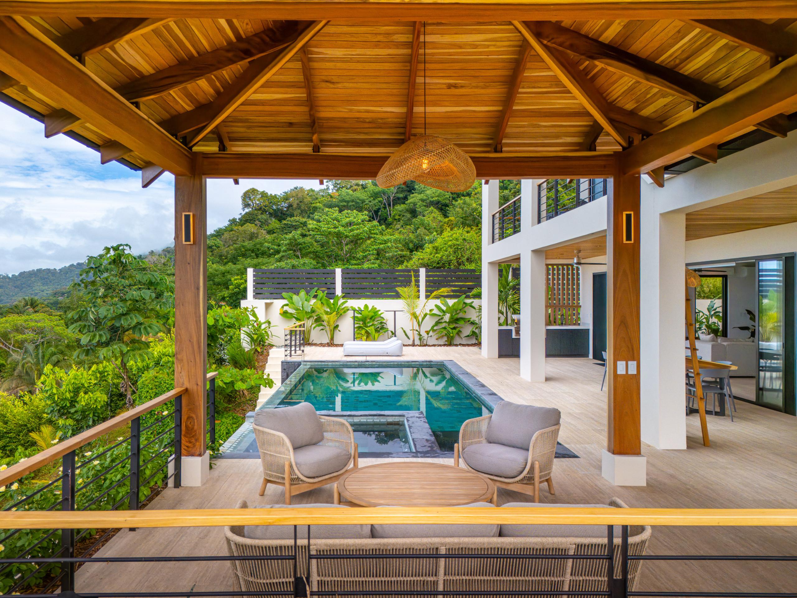 A spacious covered terrace features a high wooden ceiling and comfortable seating, offering expansive views of the adjacent infinity pool and lush jungle landscape.