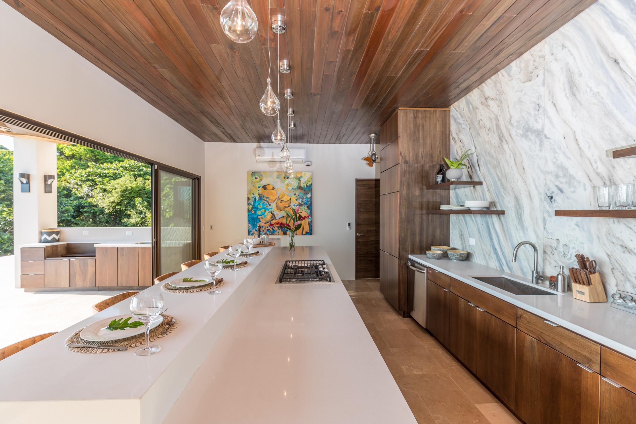 A modern open-plan kitchen features a long island with a stovetop and bar seating, a marble accent wall, warm wood ceilings, and sliding doors opening to a lush jungle view.