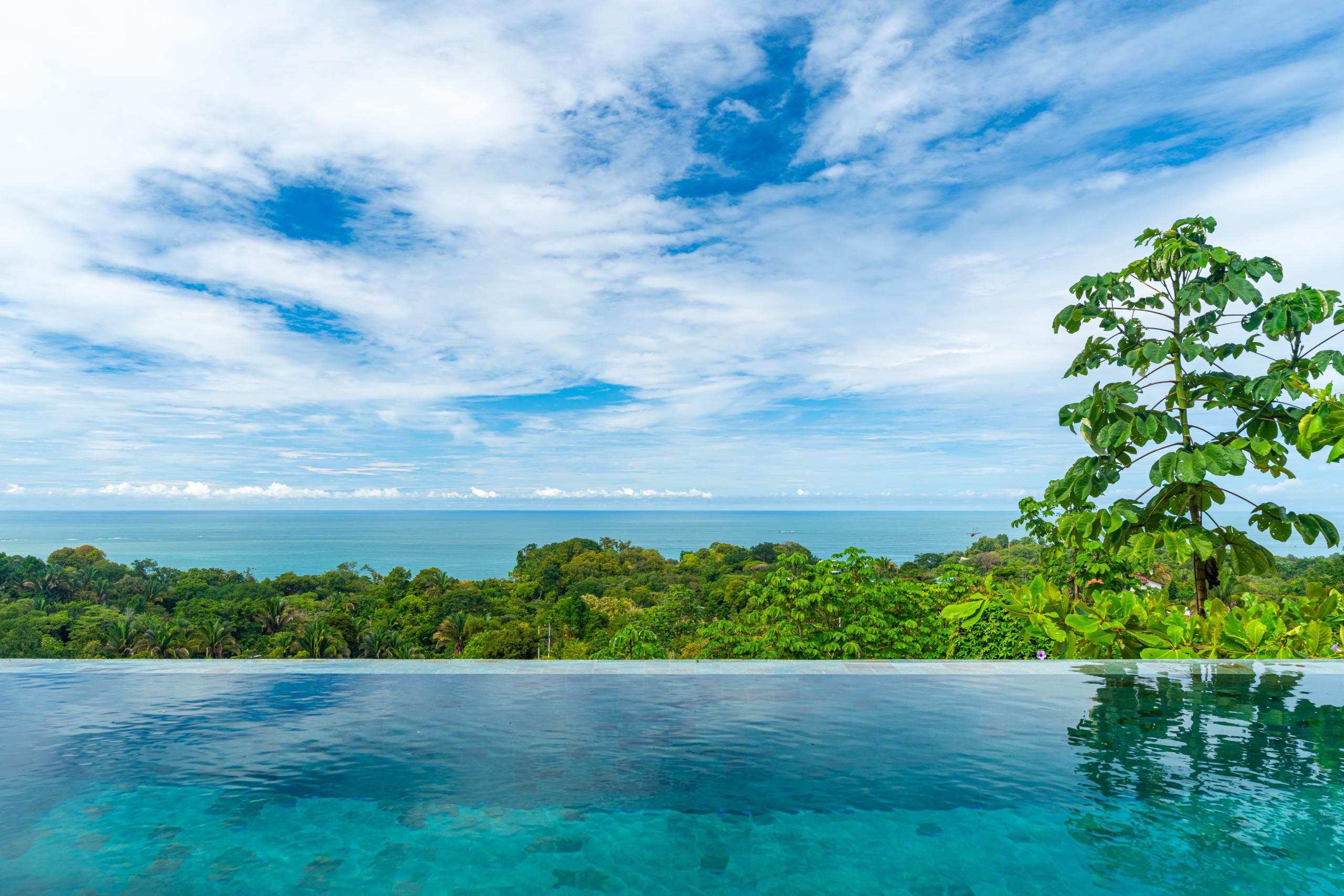 An infinity pool with clear blue water extends towards a panoramic view of the Pacific Ocean and verdant jungle beneath a cloudy sky.