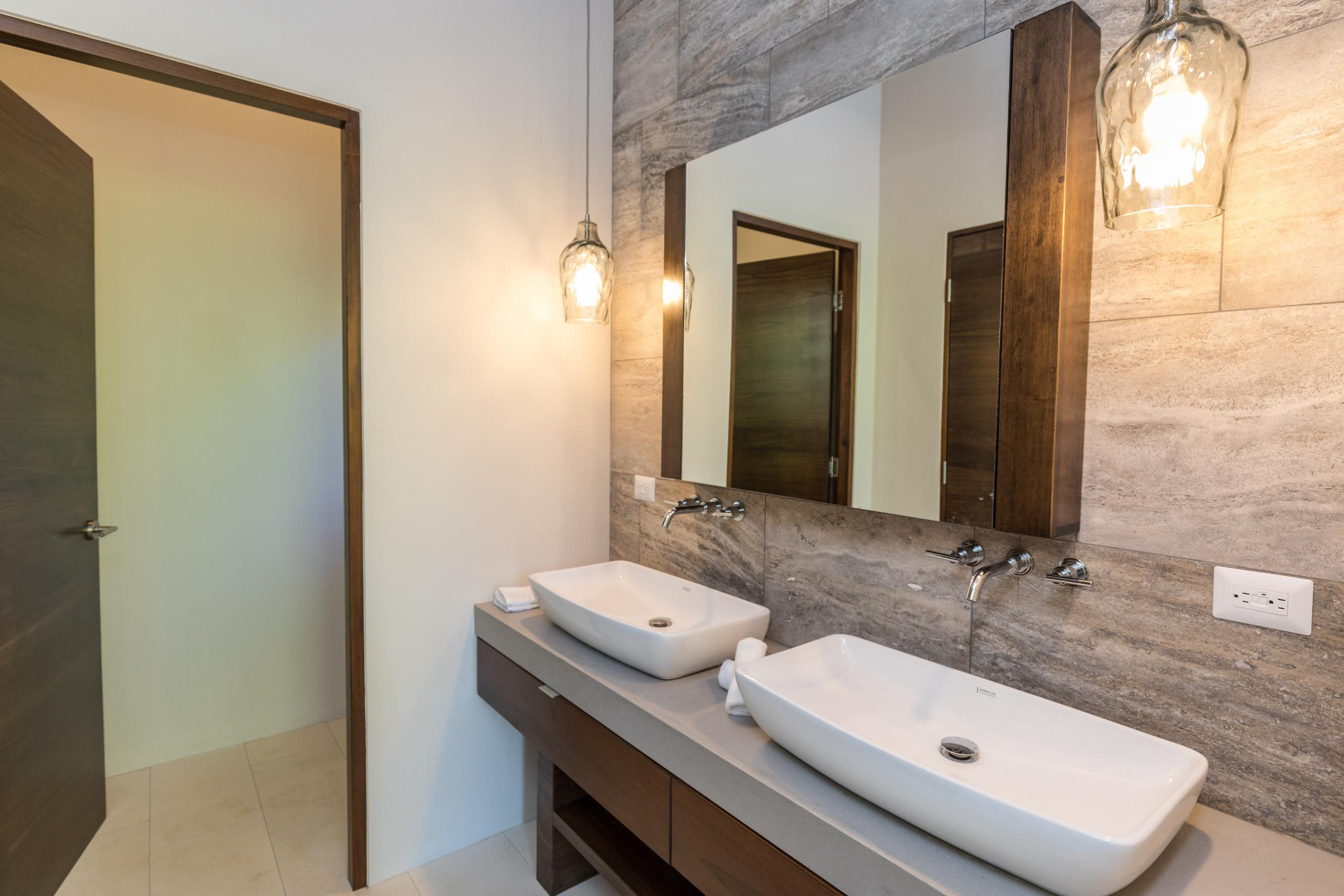 The bathroom presents a dual vanity with rectangular vessel sinks, dark wood cabinetry, and a large mirror against a textured stone wall, complemented by warm pendant and natural light.