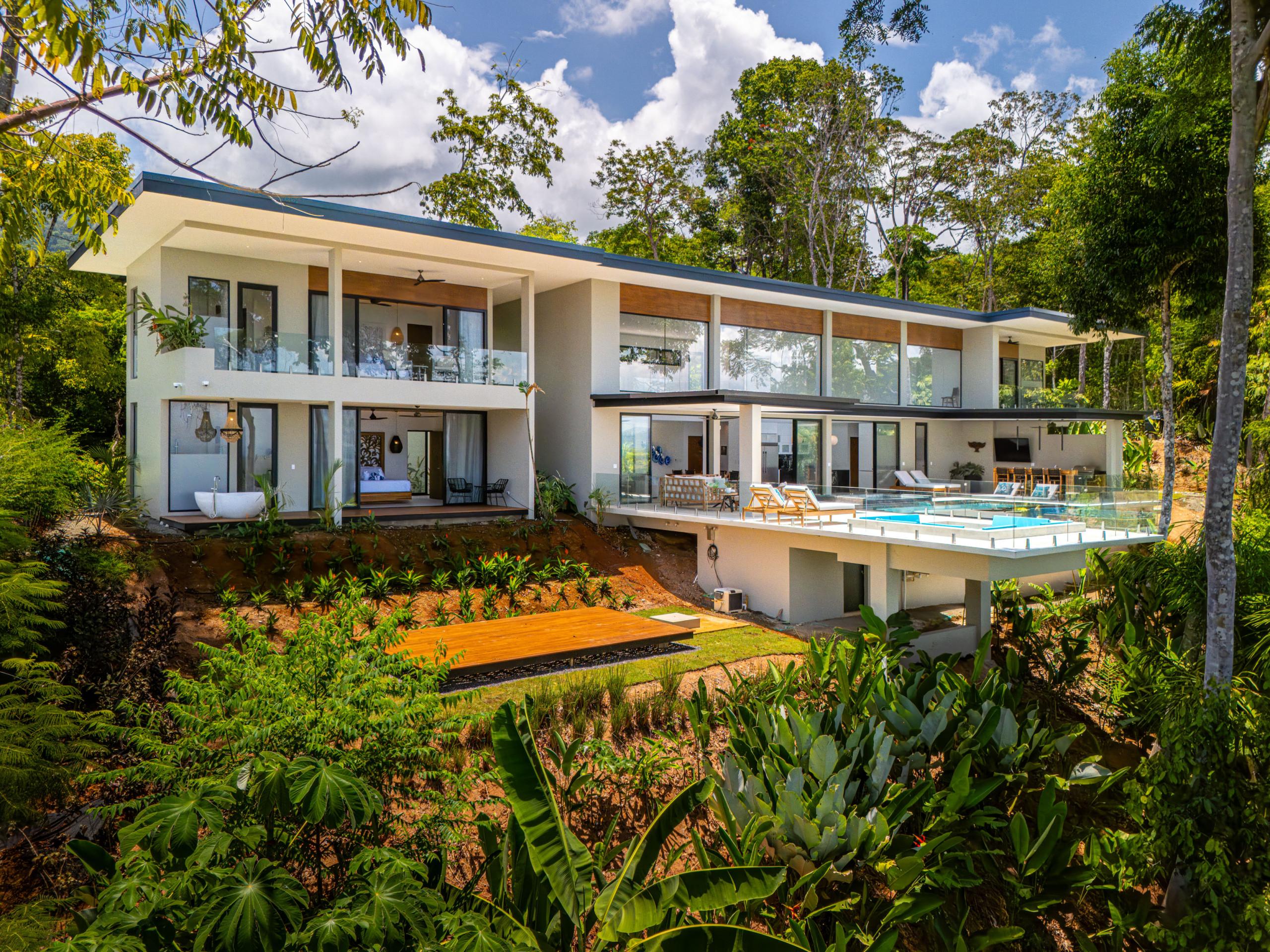 The multi-level modern villa, featuring extensive glass and open-air terraces, is immersed in a vibrant tropical landscape, offering expansive outdoor living with an infinity pool and jacuzzi overlooking distant views.