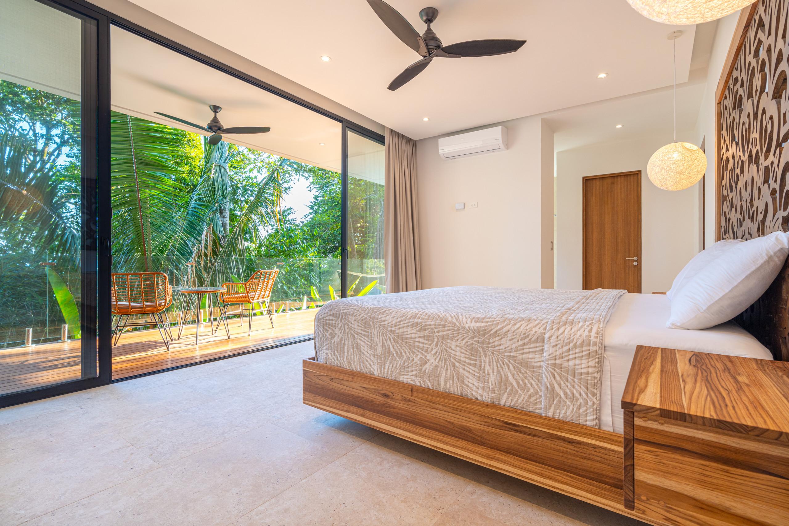 A modern bedroom with a king bed and decorative wood headboard receives natural light through full-height glass doors that open to a private balcony overlooking dense tropical foliage.