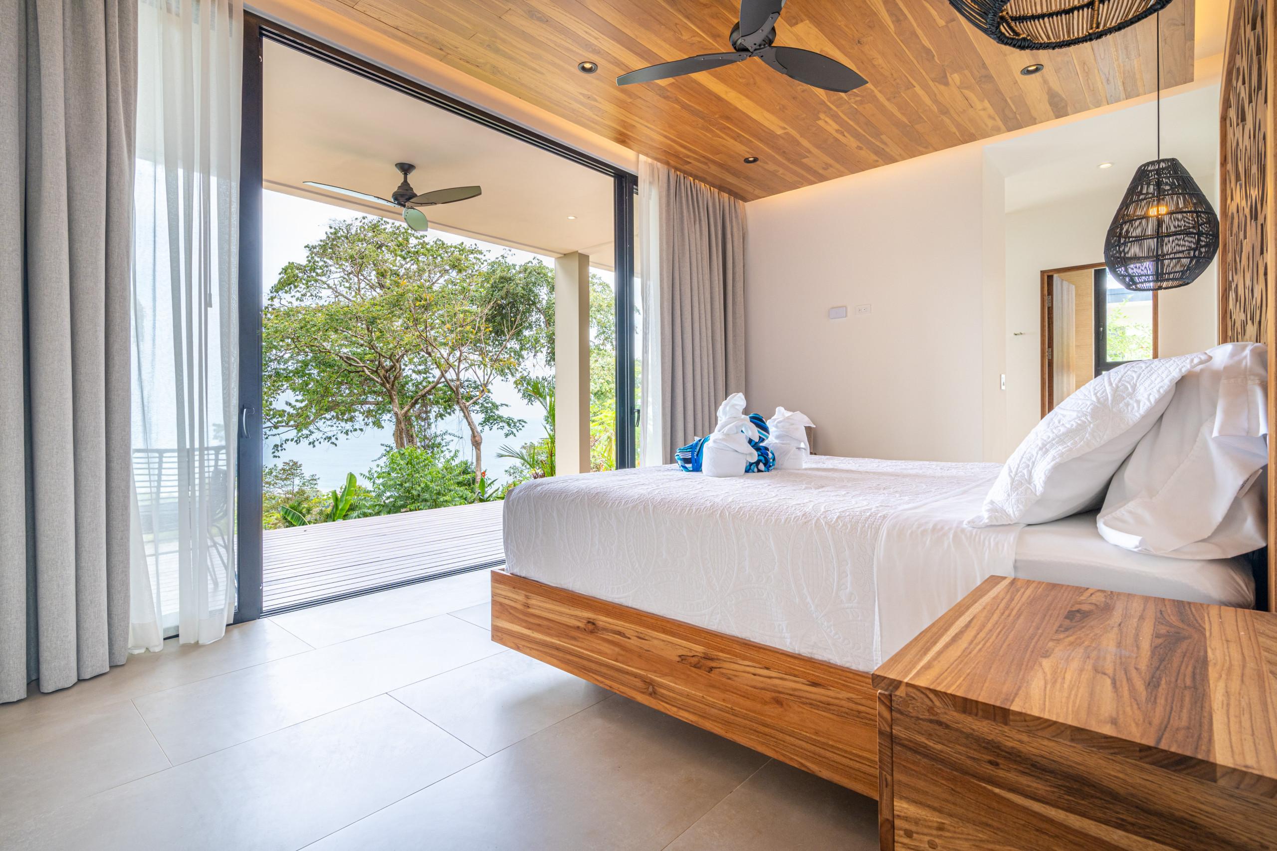 This bedroom features a king-size bed, a wooden ceiling, and full-height glass doors opening to a private terrace with tropical foliage and ocean views, allowing abundant natural light.